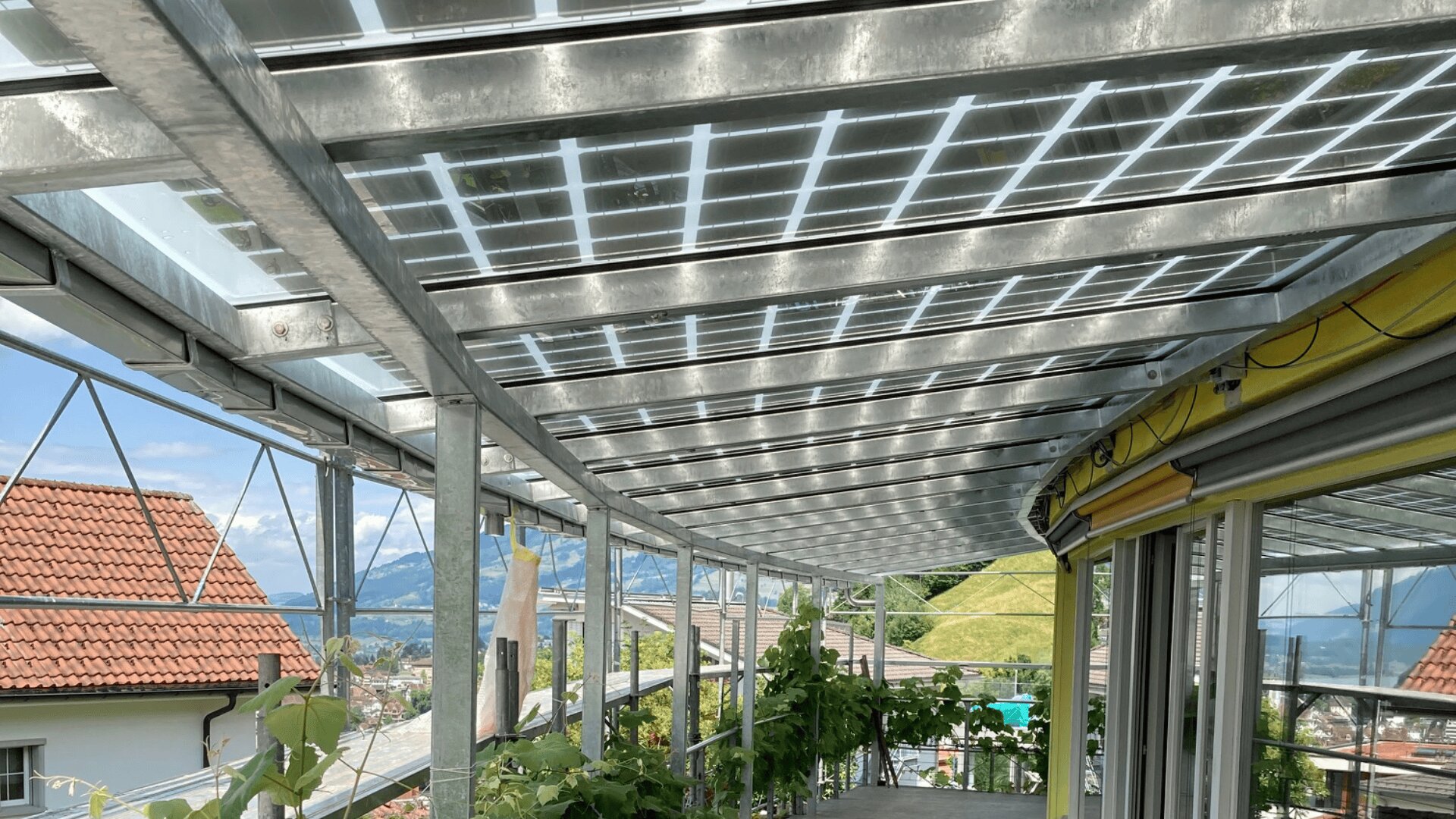 Eine überdachte Außenterrasse mit einem Glasdach mit integrierten Sonnenkollektoren, die das Licht durchlassen. Die Struktur bietet einen Blick auf Dächer, Grünflächen und ferne Berge unter einem hellen Himmel.