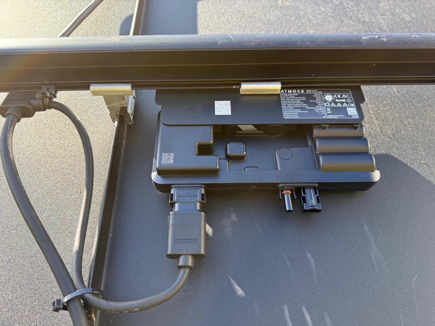 Nahaufnahme einer Anschlussdose für Solarmodule mit angeschlossenen Kabeln und Steckern, die auf der Rückseite eines Solarmoduls montiert ist. Auf der Oberfläche der Dose befinden sich Etiketten und ein QR-Code.