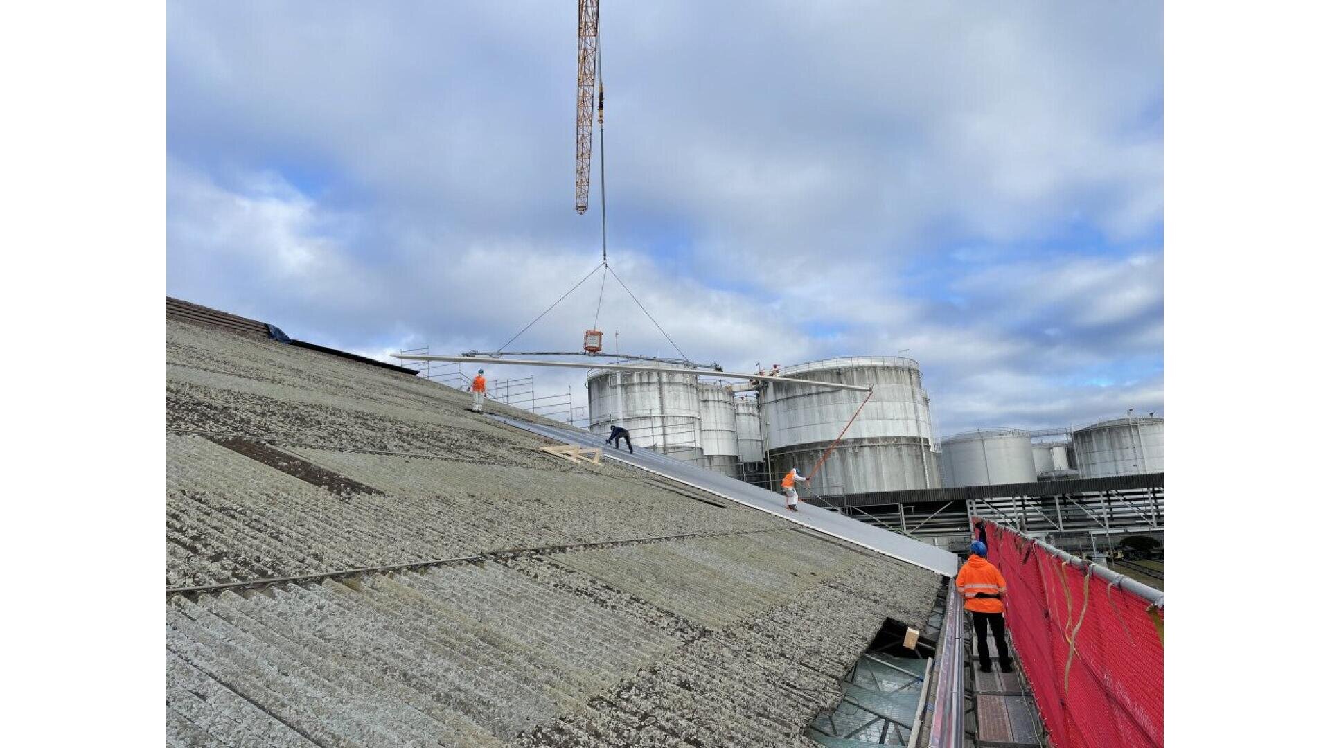 Des ouvriers en tenue de sécurité installent ou réparent un grand toit industriel en pente. Une grue soulève une poutre métallique au-dessus, avec des réservoirs de stockage de carburant et un ciel nuageux en arrière-plan. Une barrière de sécurité rouge borde le toit.