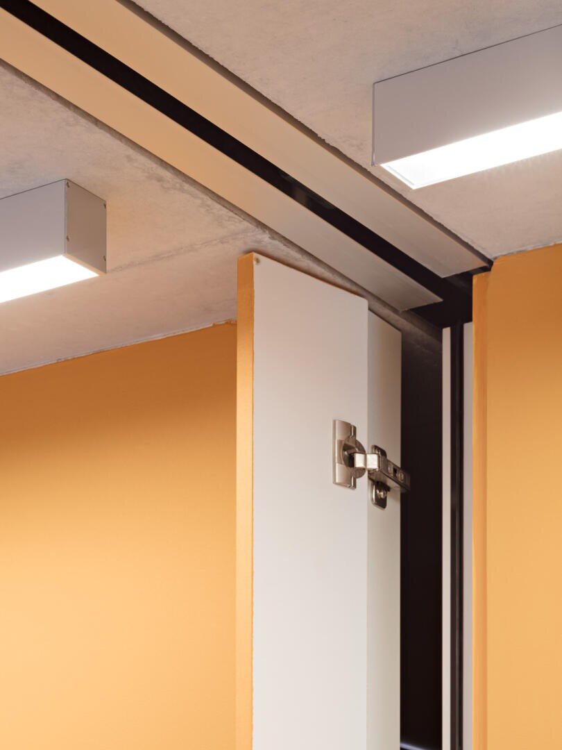 Gros plan sur une porte blanche avec une charnière métallique, partiellement ouverte, devant des murs orange et un plafond avec des luminaires rectangulaires.