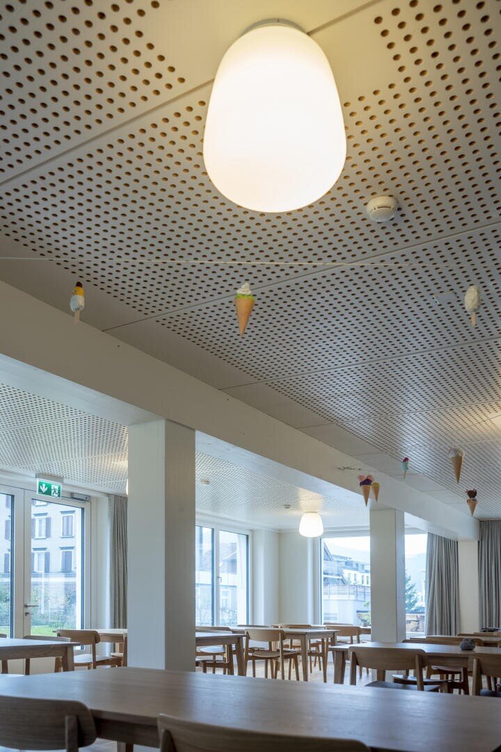 Eine moderne Cafeteria mit Holztischen und -stühlen, großen Fenstern und weißen, perforierten Decken, die mit kleinen, hängenden Eistüten verziert sind. Im Vordergrund ist eine leuchtende Deckenleuchte zu sehen.