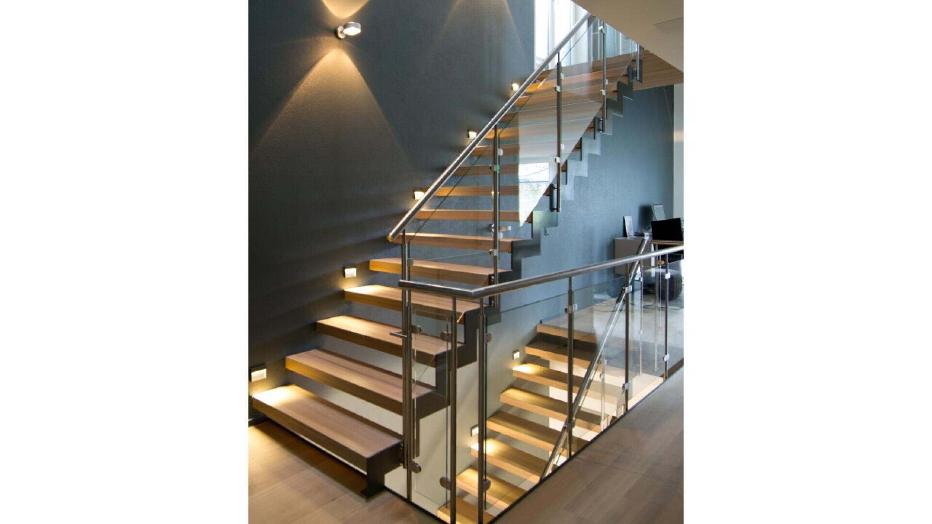 Un escalier moderne avec des marches en bois, des rampes en verre et des supports en métal est éclairé par de petites lampes le long du mur et des marches, sur un mur gris foncé dans un intérieur contemporain.