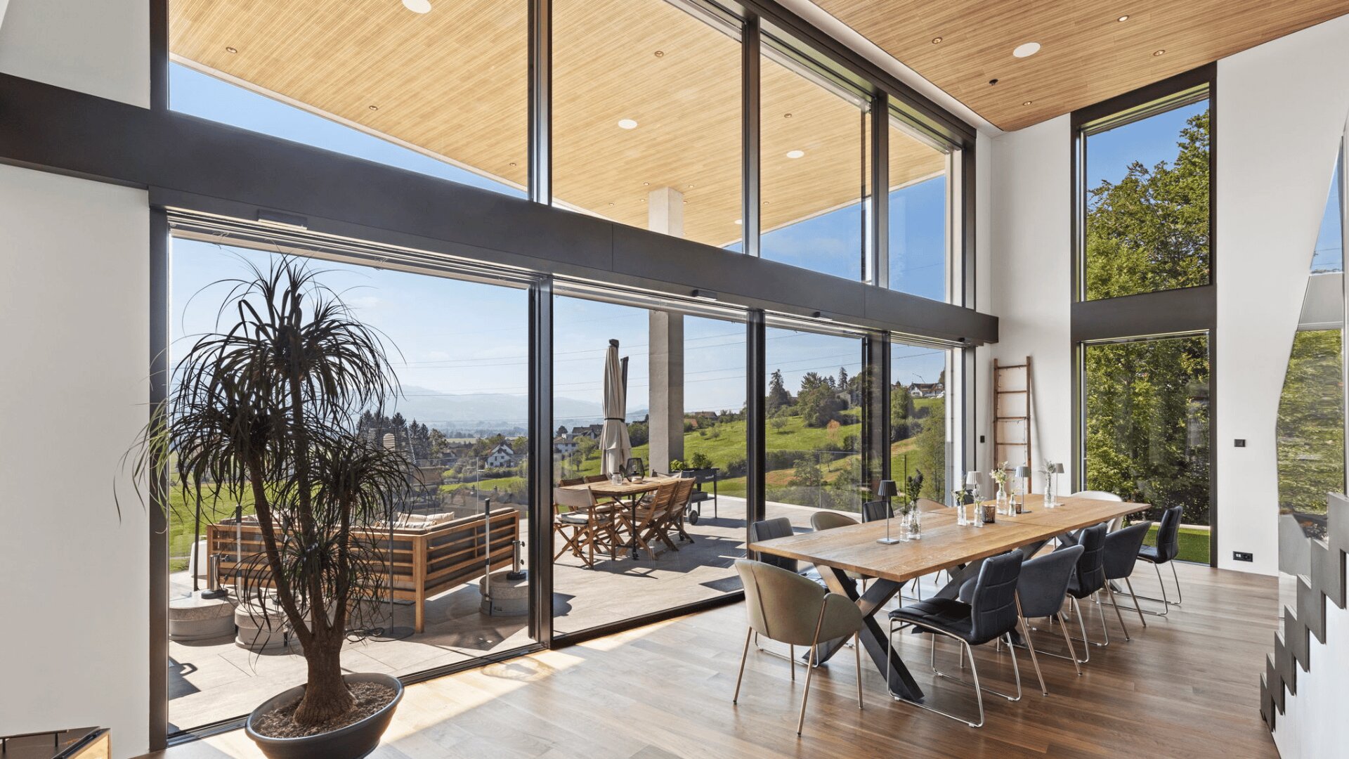Modernes Esszimmer mit raumhohen Fenstern, Holzdecke, langem Esstisch mit Stühlen, Zimmerpflanze und Sitzgelegenheiten im Freien mit Blick auf eine malerische Landschaft mit Bäumen und Hügeln.