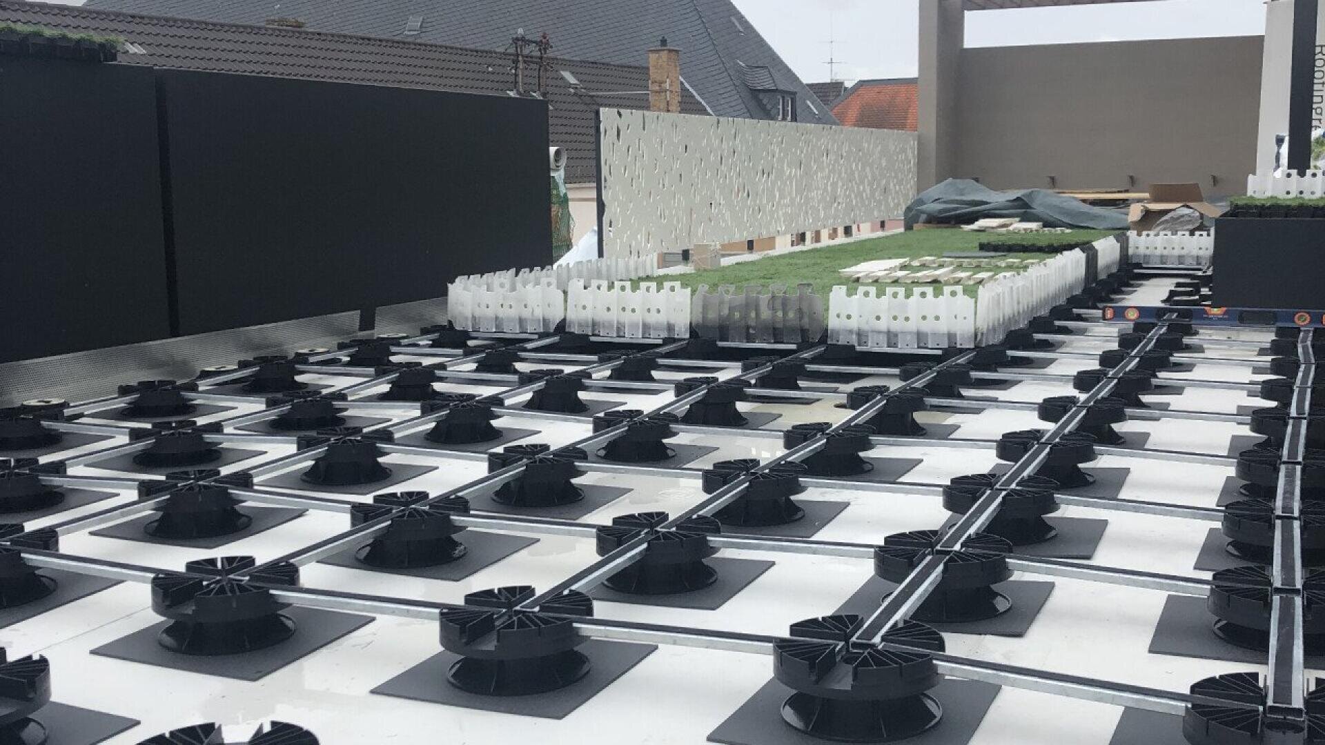Une installation sur le toit présente des socles ajustables noirs supportant des tuiles blanches, avec des bacs de toiture verts et des matériaux de construction empilés à l'arrière-plan sur fond de ciel couvert.