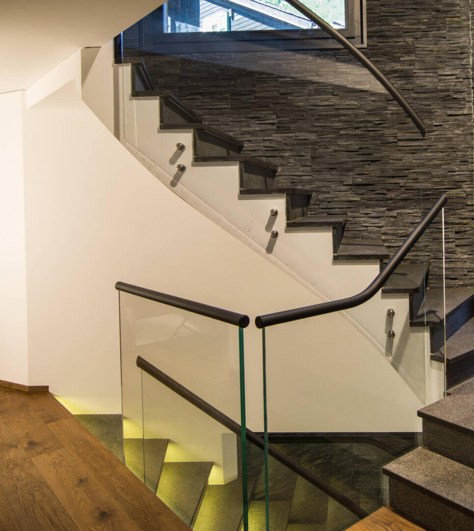 Moderne Treppe mit Glas- und Metallgeländer, Akzentwand aus Stein und Holzfußboden. Warme Beleuchtung beleuchtet die Stufen und die untere Wand und schafft eine moderne und stilvolle Inneneinrichtung.