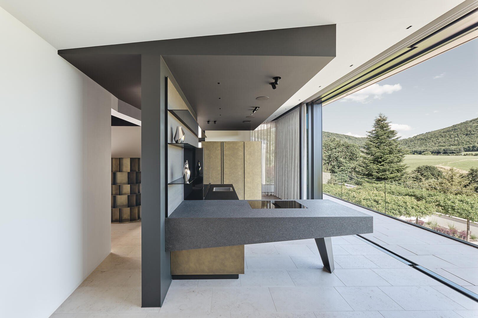 Une cuisine moderne avec des armoires grises minimalistes, un plan de travail lisse et flottant, des étagères ouvertes avec des articles de décoration et de grandes portes coulissantes en verre qui donnent sur un paysage pittoresque de collines et d'arbres verdoyants.