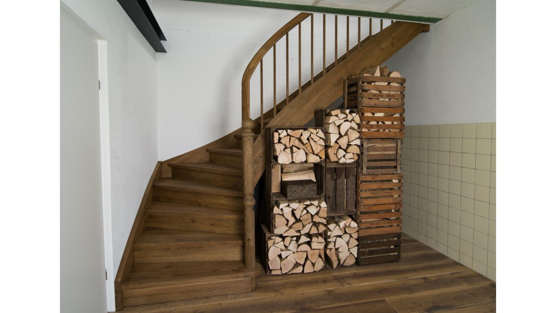 Un escalier en bois avec une rampe incurvée. Sous l'escalier, des caisses en bois sont empilées et remplies de bois de chauffage bien rangé. Le sol et l'escalier sont en bois, et un mur est carrelé.