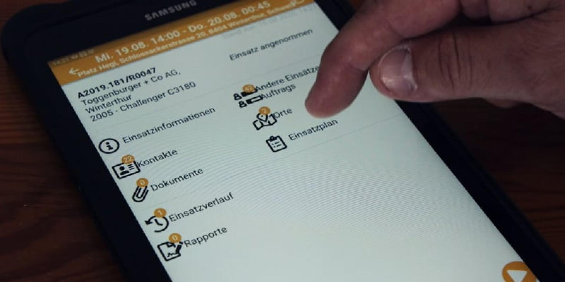 Eine Hand interagiert mit einem Samsung-Smartphone, das eine deutschsprachige Notfall- oder Einsatzmanagement-App mit Menüoptionen wie Einsatzinformationen, Kontakte, Dokumente und mehr anzeigt.