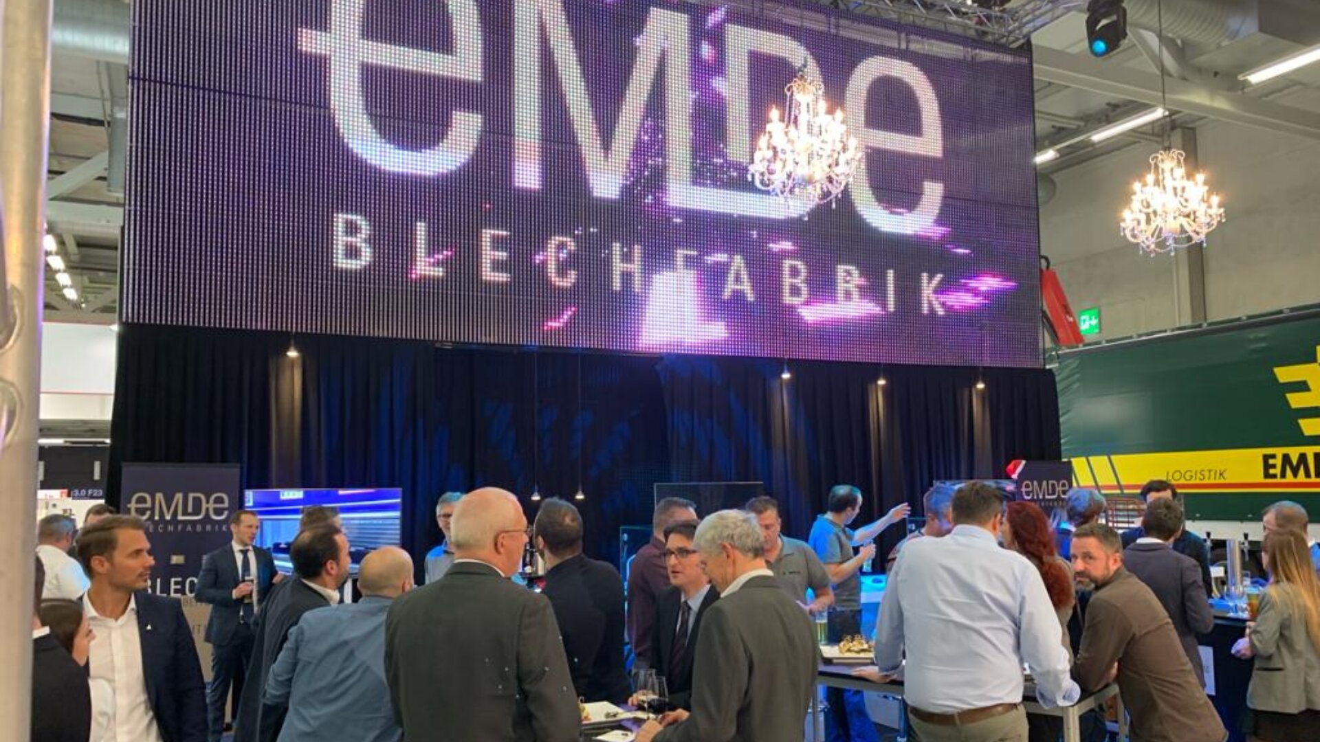 Eine Gruppe von Menschen in Geschäftskleidung versammelt sich bei einer Networking-Veranstaltung oder einer Fachmesse. Große Bildschirme zeigen das Wort EMDE BLECHFABRIK über der Menge an, und von der Decke hängen Kronleuchter.