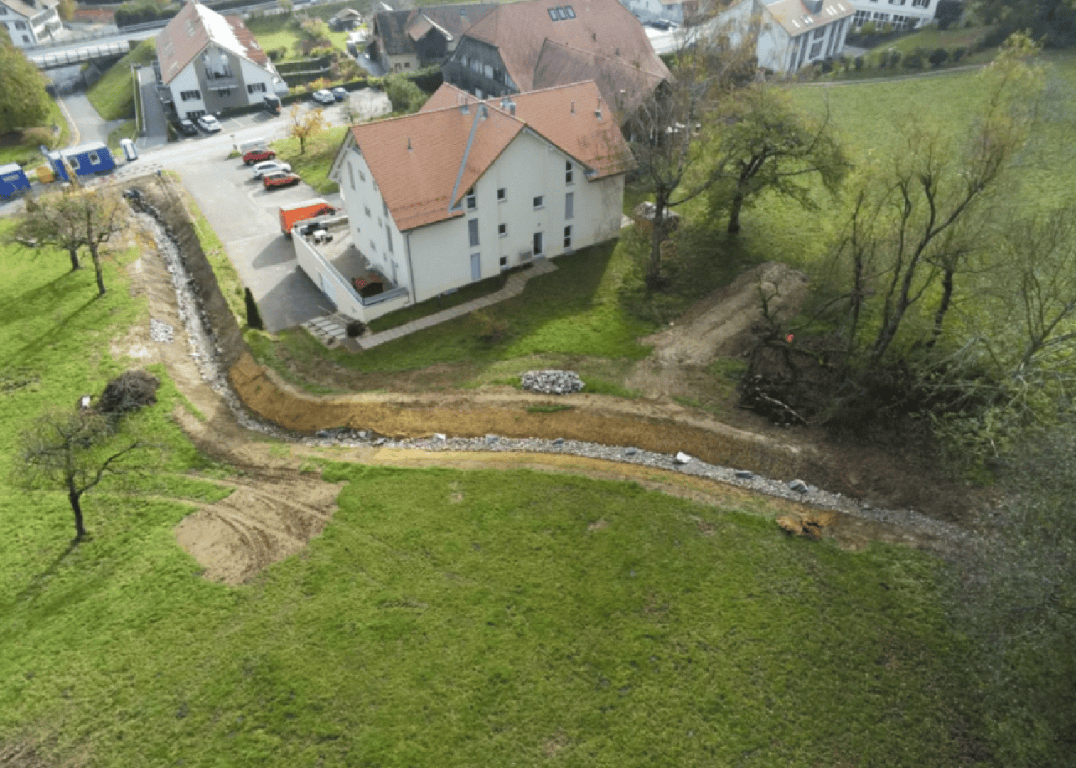 Luftaufnahme eines hellen Gebäudes neben einem gekrümmten, kiesgesäumten und im Bau befindlichen Weg oder Wasserlauf, umgeben von grünem Gras, Bäumen und Nachbarhäusern in einem Wohngebiet.