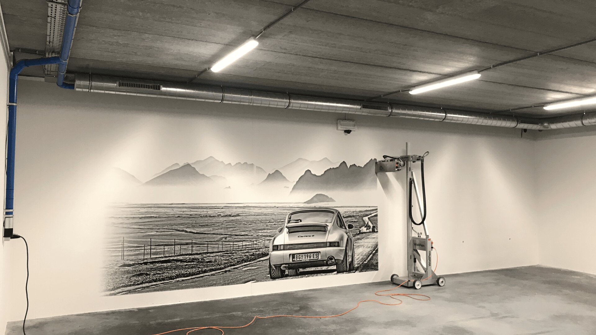 Dans un garage dont le plafond et le sol sont en béton, une peinture murale en noir et blanc représente une voiture de collection roulant vers les montagnes. Un appareil de nettoyage muni d'un cordon orange est fixé sur le mur à droite de la peinture murale.