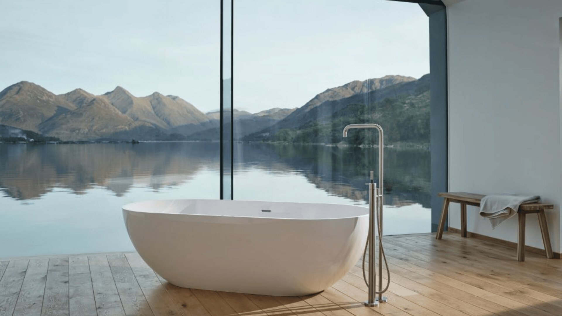 Eine moderne weiße Badewanne steht auf einem Holzfußboden in einem minimalistischen Badezimmer mit bodentiefen Fenstern, die einen atemberaubenden Blick auf einen ruhigen See und ferne Berge unter einem klaren Himmel bieten. Eine Holzbank mit Handtüchern steht in der Nähe.
