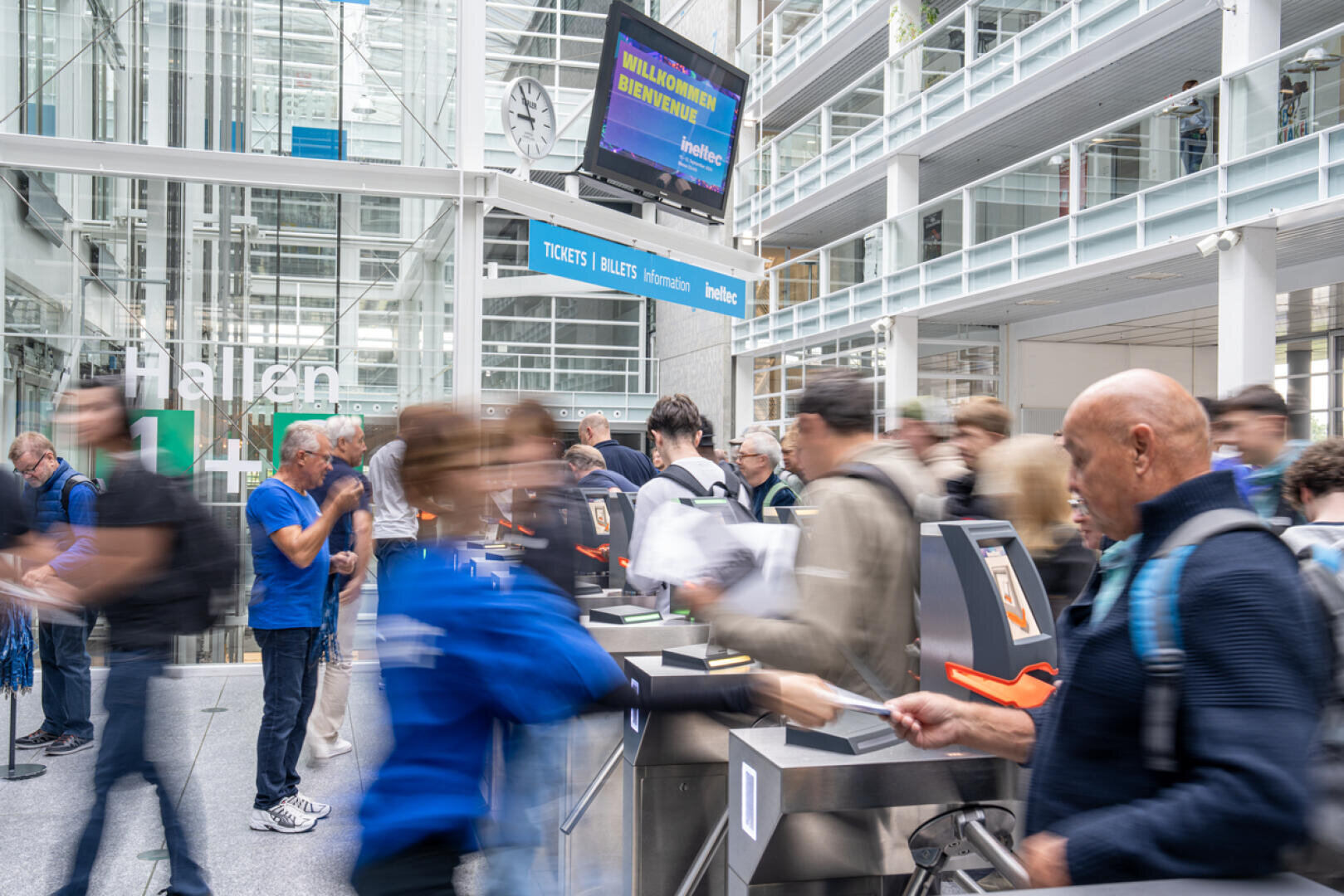 Eine belebte Ticketkontrolle in einem Innenbereich, an der Menschen durch Drehkreuze gehen. Einige Mitarbeiter tragen blaue Hemden und stehen den Besuchern zur Seite. Über ihnen ist ein großes Schild mit der Aufschrift „Willkommen“ zu sehen, und im Hintergrund sind Glaswände und moderne Architektur zu erkennen.