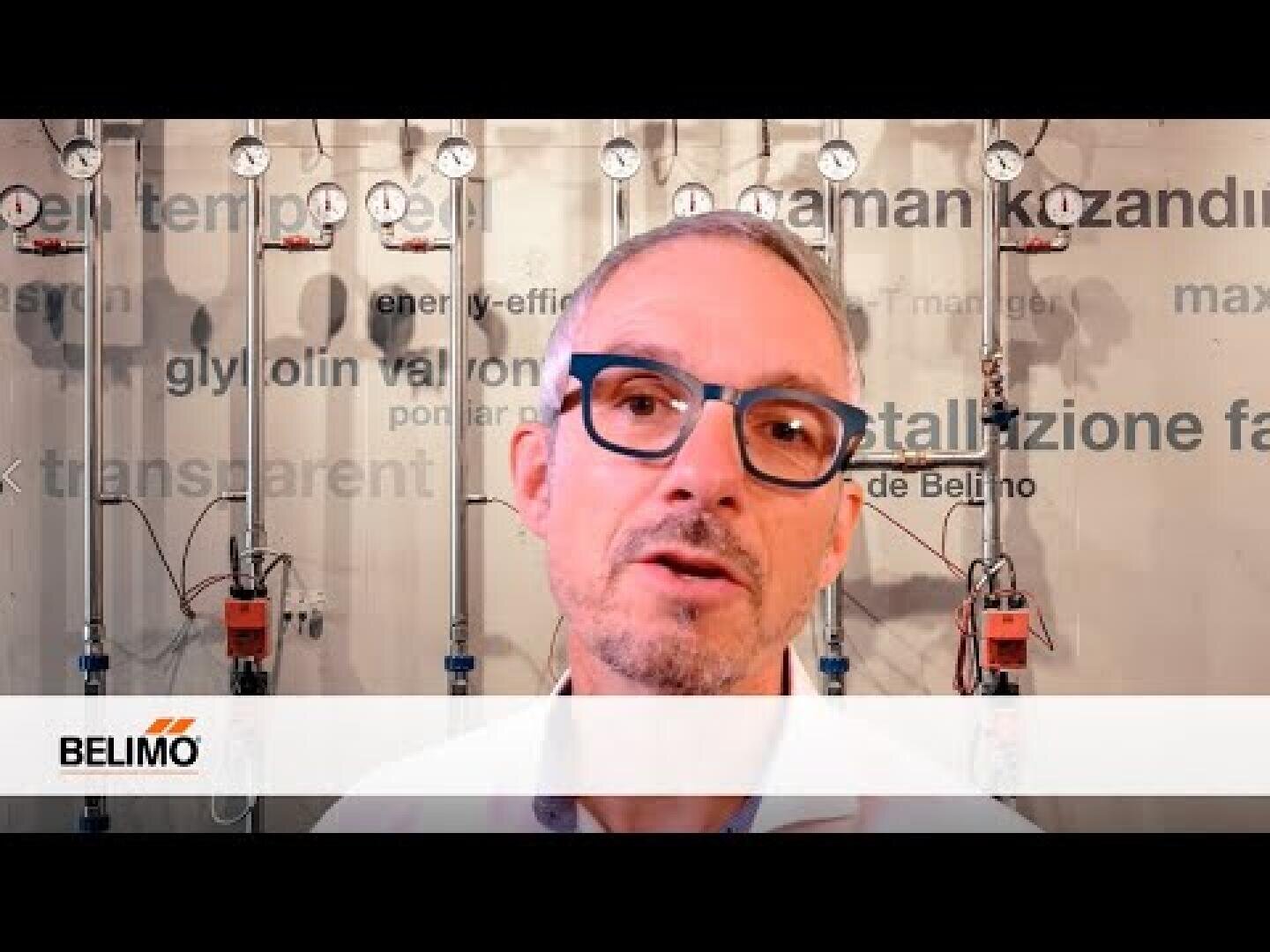 Ein Mann mit kurzen Haaren und Brille steht vor einer Wand mit Messgeräten, Rohren und dem Belimo-Logo. An der Wand hinter ihm sind Wörter zu sehen, die mit Technik und Energieeffizienz zu tun haben.