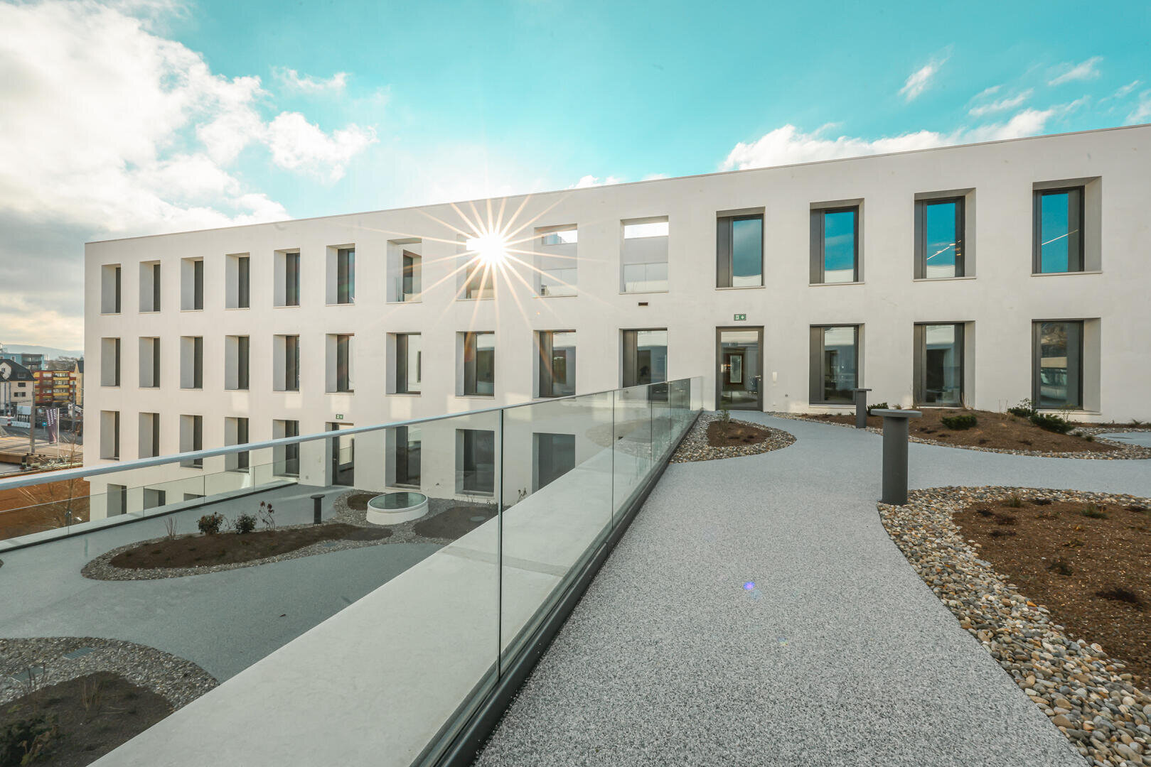 Modernes weißes Gebäude mit großen Fenstern, einem Glasgeländer und einem begrünten Gehweg im Vordergrund. Die Sonne scheint hell und erzeugt einen Lens-Flare-Effekt auf der Fassade des Gebäudes.
