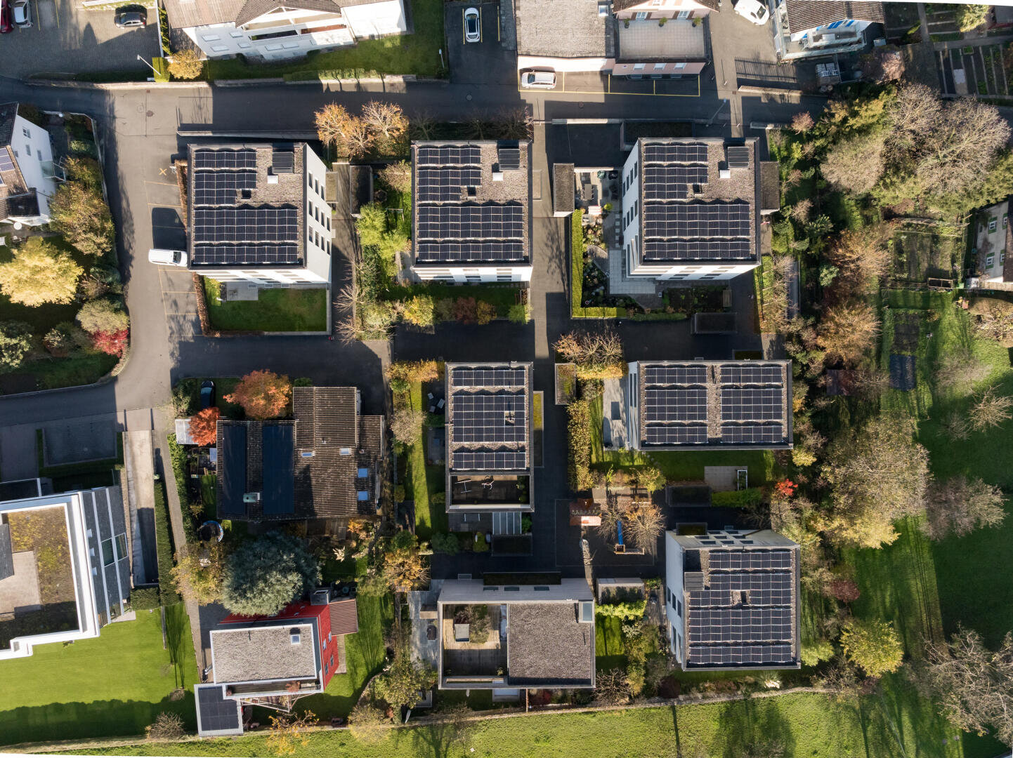 Luftaufnahme eines Wohngebiets mit mehreren modernen Gebäuden, die mit Solarzellen auf dem Dach ausgestattet sind, umgeben von grünen Rasenflächen, Bäumen und Straßen. Das Layout ist geordnet und geometrisch.