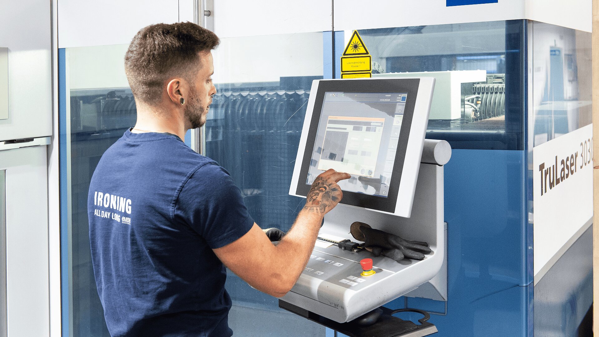 Ein Mann in einem blauen IRONING-T-Shirt bedient ein Touchscreen-Bedienfeld in einer modernen Industrieanlage, während im Hintergrund Maschinen und Sicherheitsschilder zu sehen sind.