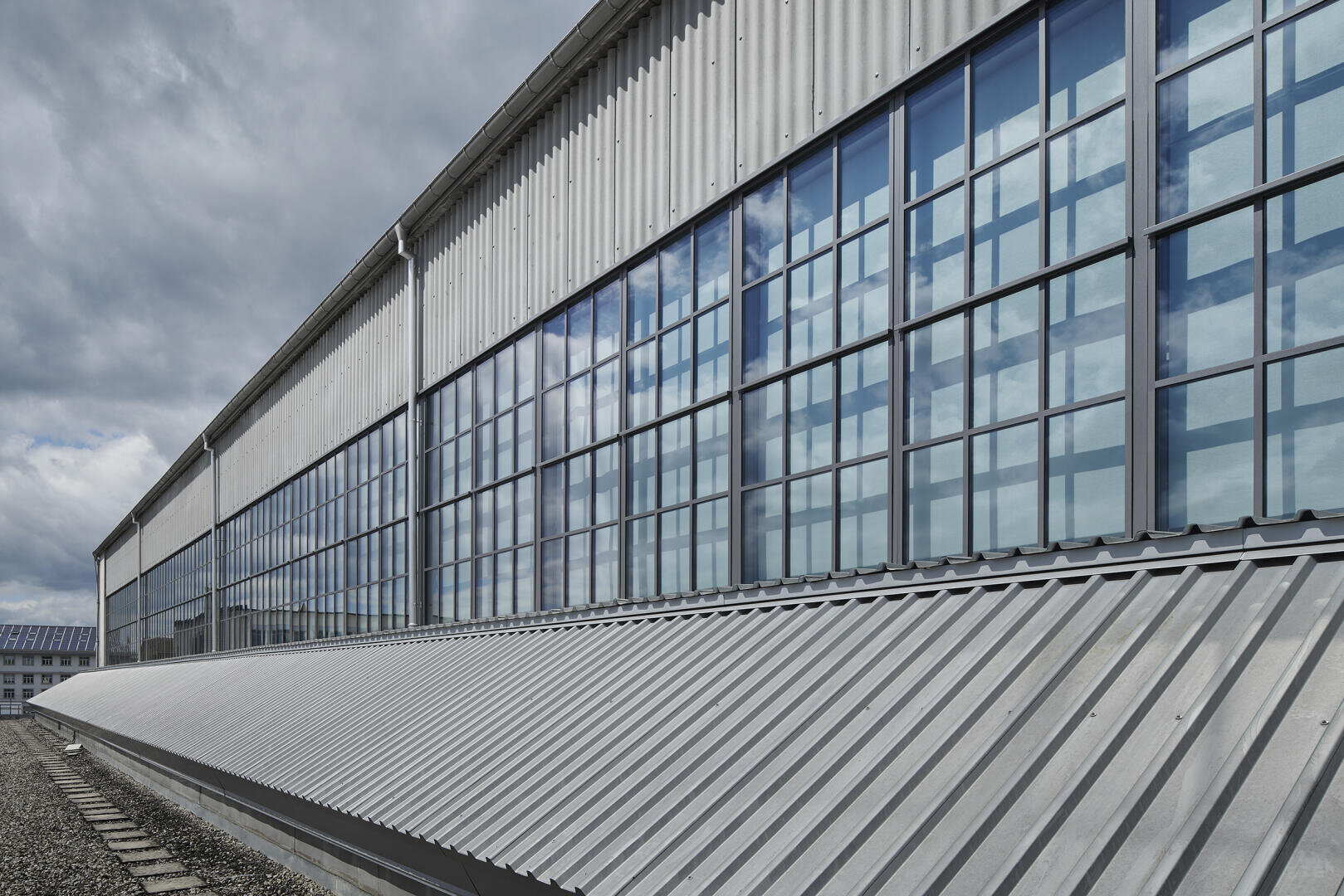 Ein großes Industriegebäude mit hohen, rechteckigen Fenstern, in denen sich der bewölkte Himmel spiegelt, und einem schrägen Metalldach, von außen betrachtet.
