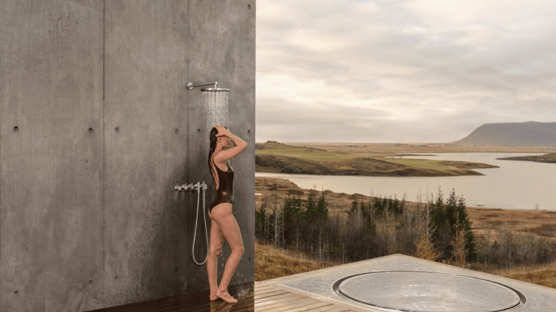 Eine Frau im Badeanzug duscht im Freien vor einer Betonwand mit einer malerischen Kulisse aus Feldern, einem Fluss und fernen Hügeln unter einem bewölkten Himmel. Im Vordergrund ist ein Whirlpool zu sehen.