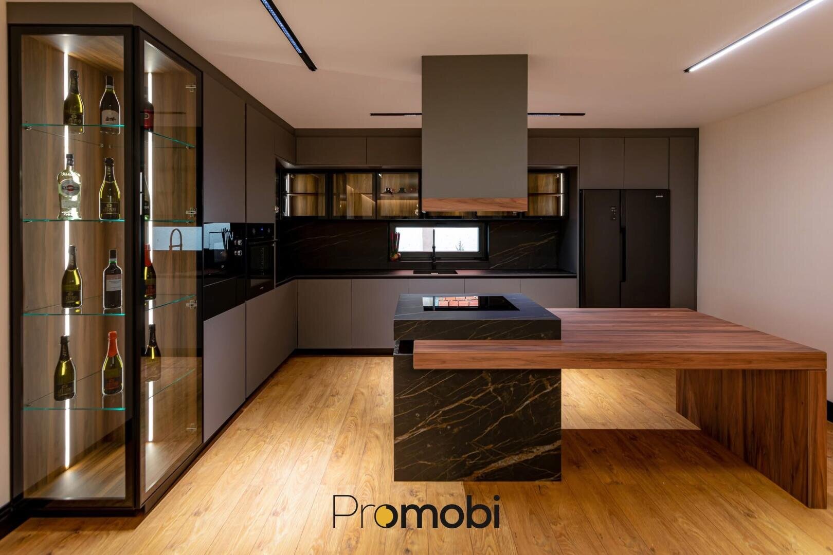 Cuisine moderne avec des armoires élégantes, une vitrine de bouteilles, un îlot noir avec une extension de comptoir en bois, des appareils encastrés, un réfrigérateur noir et un sol en bois. Le mot Promobi apparaît en bas au centre.