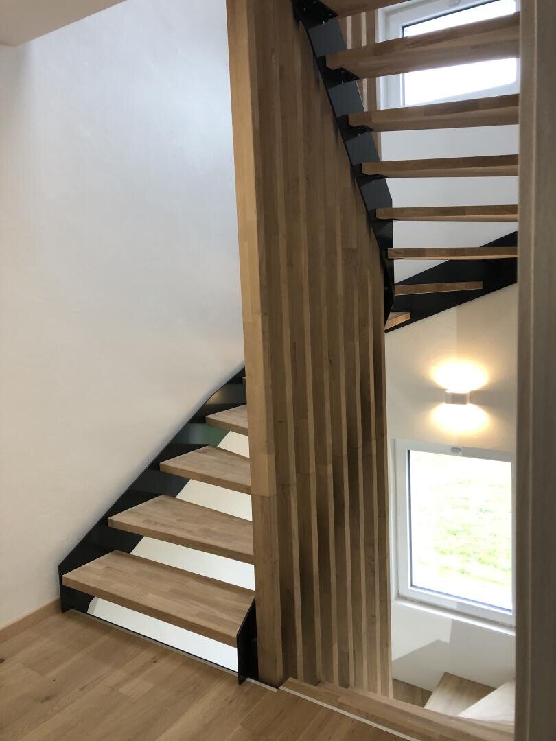 Un escalier moderne en bois avec des contremarches ouvertes et des supports en métal noir, avec des lattes verticales en bois en guise de séparation partielle. Une fenêtre et une lampe murale apportent de la lumière naturelle et artificielle à l'espace.