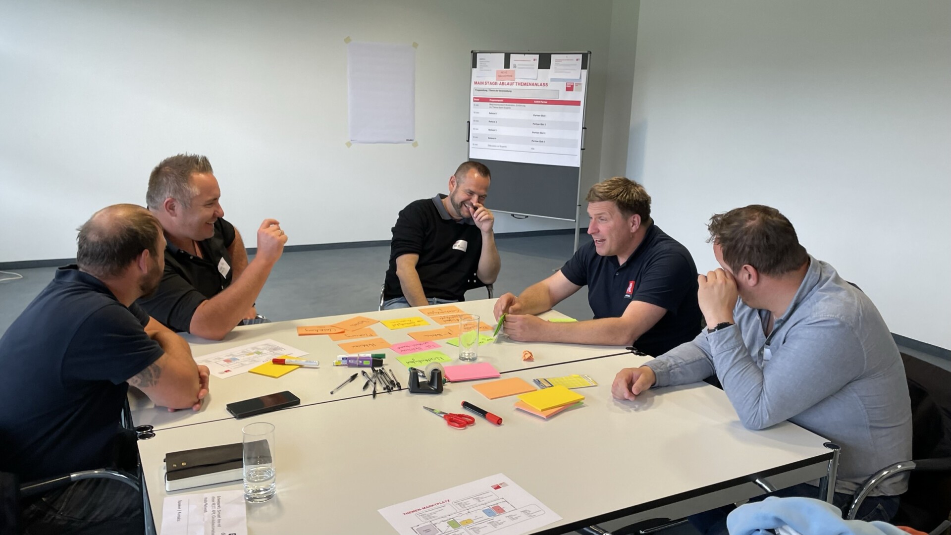 Fünf Männer sitzen um einen Tisch und diskutieren. Vor ihnen liegen bunte Haftnotizen, Stifte und Papiere ausgebreitet. Im Hintergrund sind ein Flipchart und ein großes Blatt mit Text zu sehen.