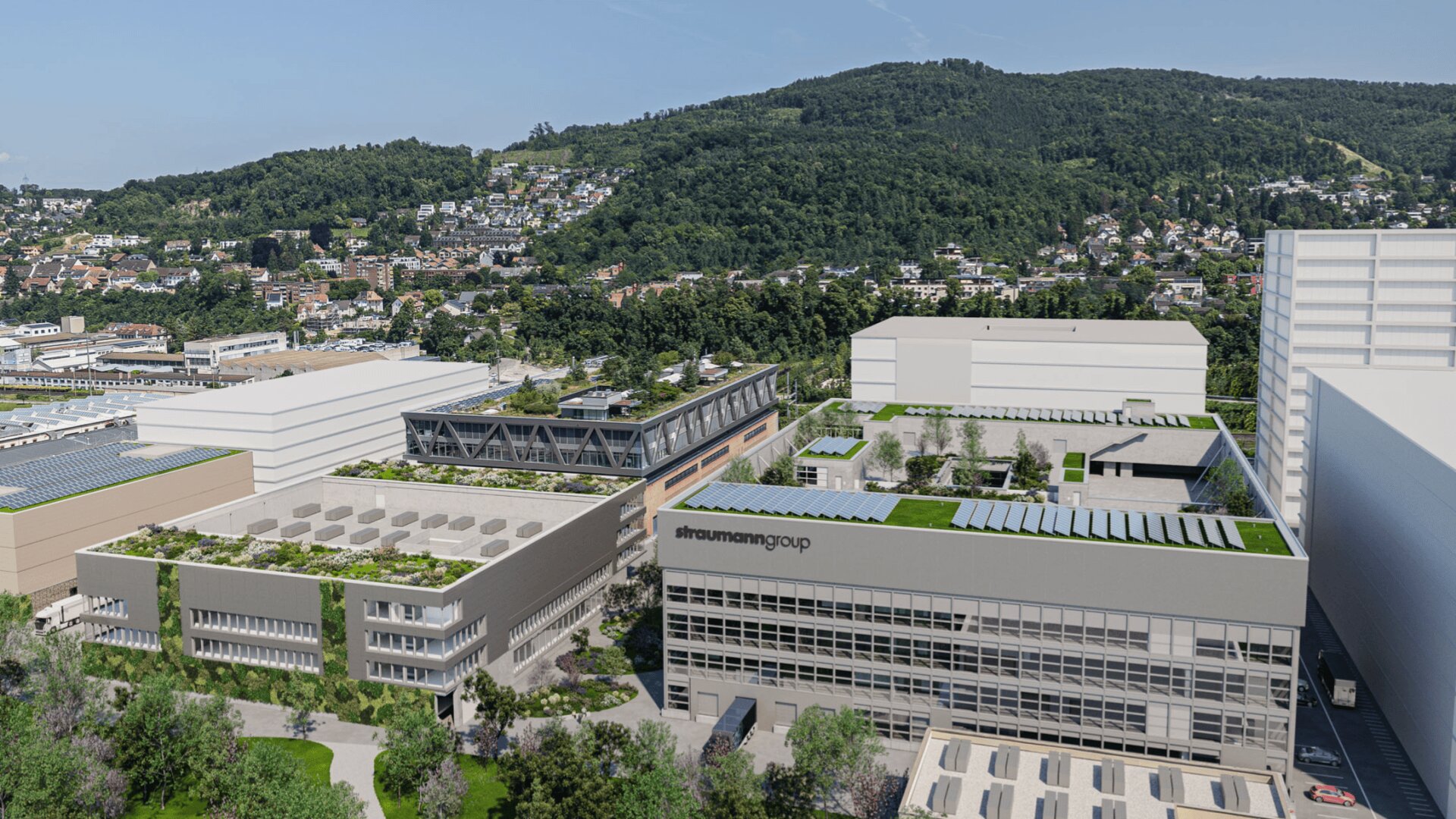Ein moderner Industriekomplex mit mehreren großen, rechteckigen Gebäuden mit Dachgärten, umgeben von Grünflächen und Bäumen. Im Hintergrund reihen sich Häuser an einem grünen Hügel unter einem klaren Himmel.