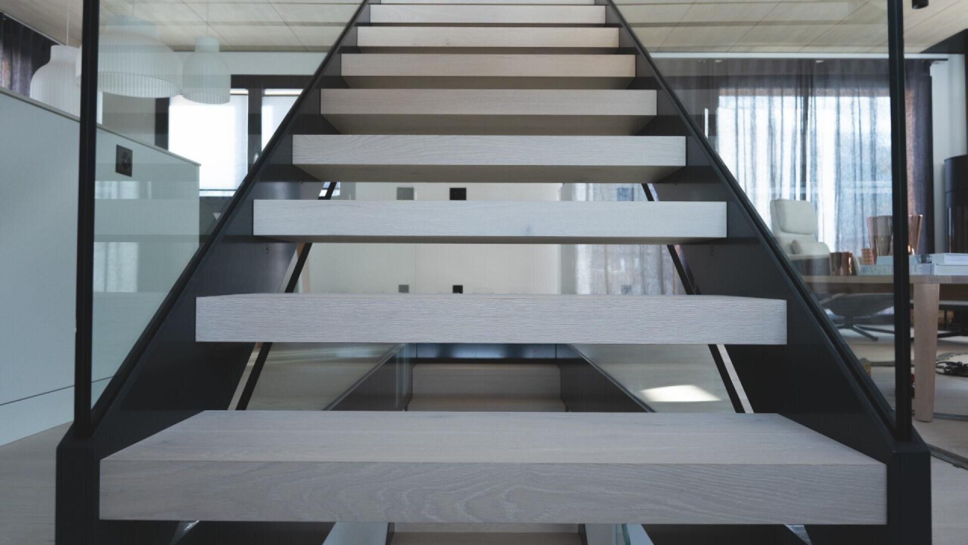 Vue rapprochée d'un escalier moderne à volée ouverte avec des marches en bois clair et des supports en métal noir dans un intérieur contemporain avec de grandes fenêtres et des garde-corps en verre.