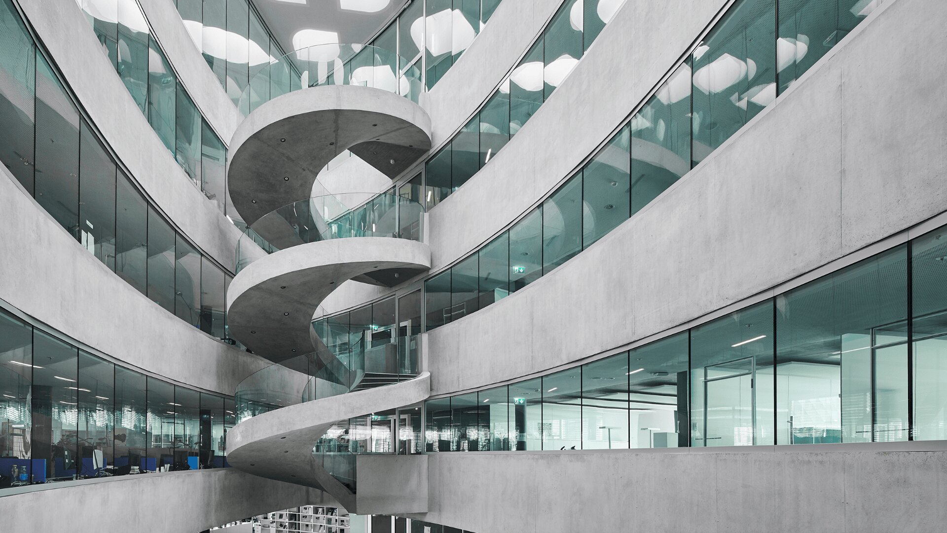 Ein modernes Gebäudeinneres mit geschwungenen Betonwänden, Glaspaneelen und einer auffälligen Wendeltreppe, die mehrere Stockwerke miteinander verbindet und ein elegantes und offenes architektonisches Design schafft.
