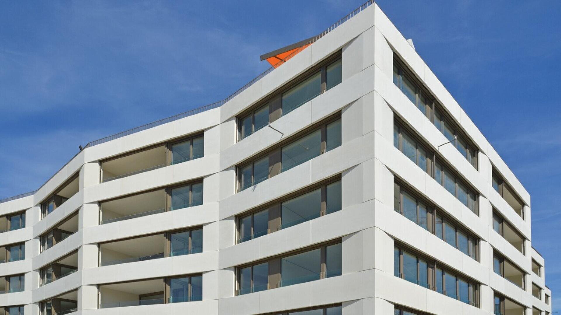 Immeuble moderne d'appartements blancs avec de grands balcons en verre et une architecture angulaire, sur fond de ciel bleu clair. L'immeuble comporte plusieurs étages et un angle aigu au premier plan.