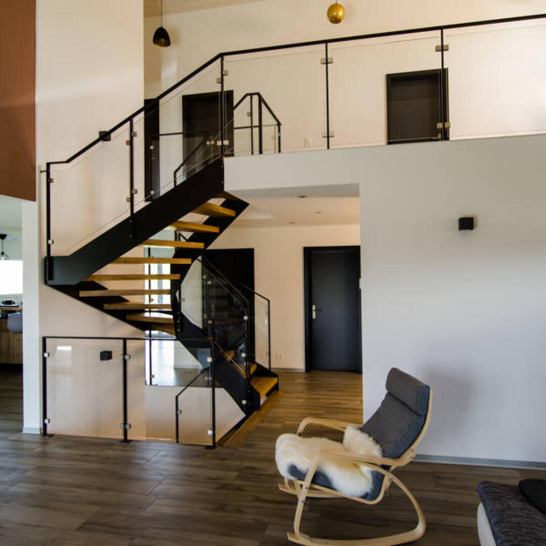 Modernes Interieur mit Holzboden, schlichter schwarzer Treppe mit Glasgeländer, weißen Wänden und einem Schaukelstuhl mit Fellüberwurf in der Nähe der Treppe.