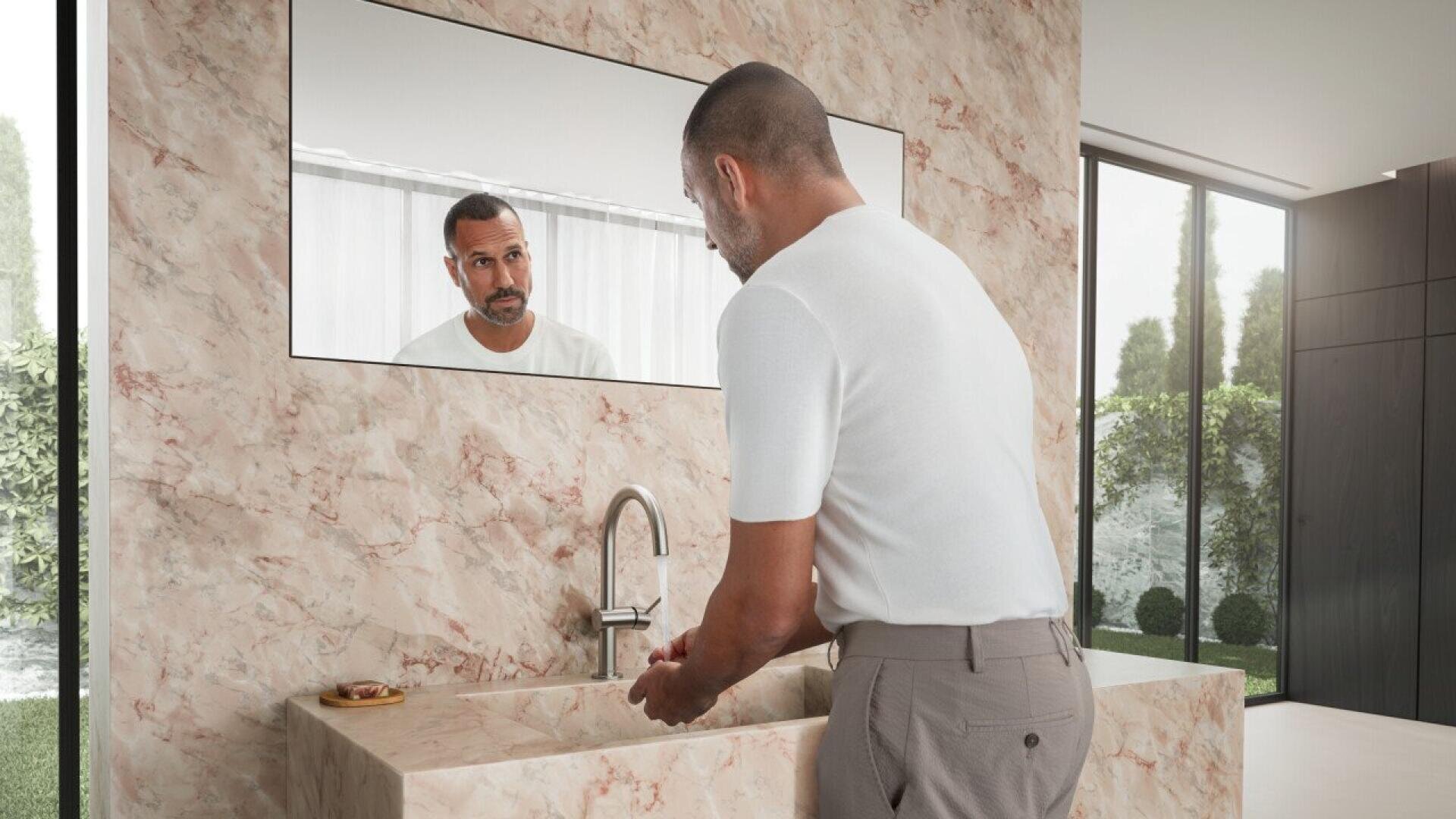 Un homme en chemise blanche se lave les mains devant un lavabo en marbre beige, en se regardant dans un grand miroir, dans une salle de bains moderne avec de grandes fenêtres et une vue sur le jardin.