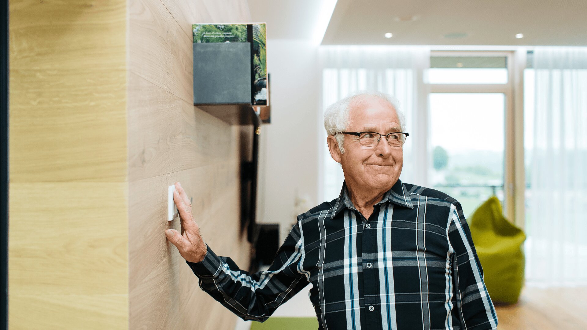 Ein älterer Mann mit Brille und kariertem Hemd steht in einem Haus, lächelt und schaltet einen Lichtschalter an einer Holzwand aus. Helles Tageslicht fällt durch große Fenster im Hintergrund.