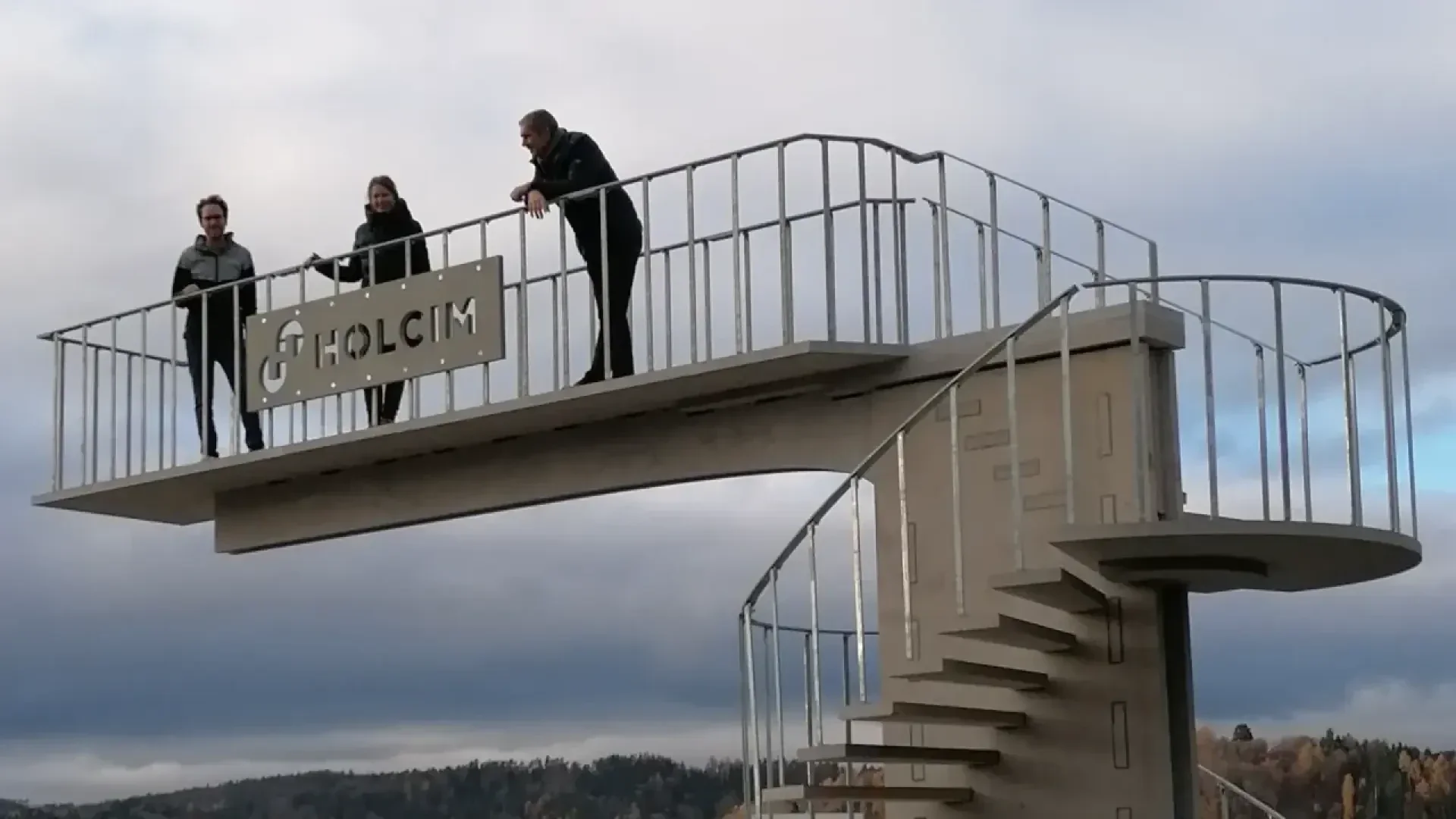 Drei Personen stehen auf einem hohen Sprungturm mit einer Wendeltreppe und einem Holcim-Schild und blicken auf eine Landschaft mit Bäumen unter einem bewölkten Himmel.
