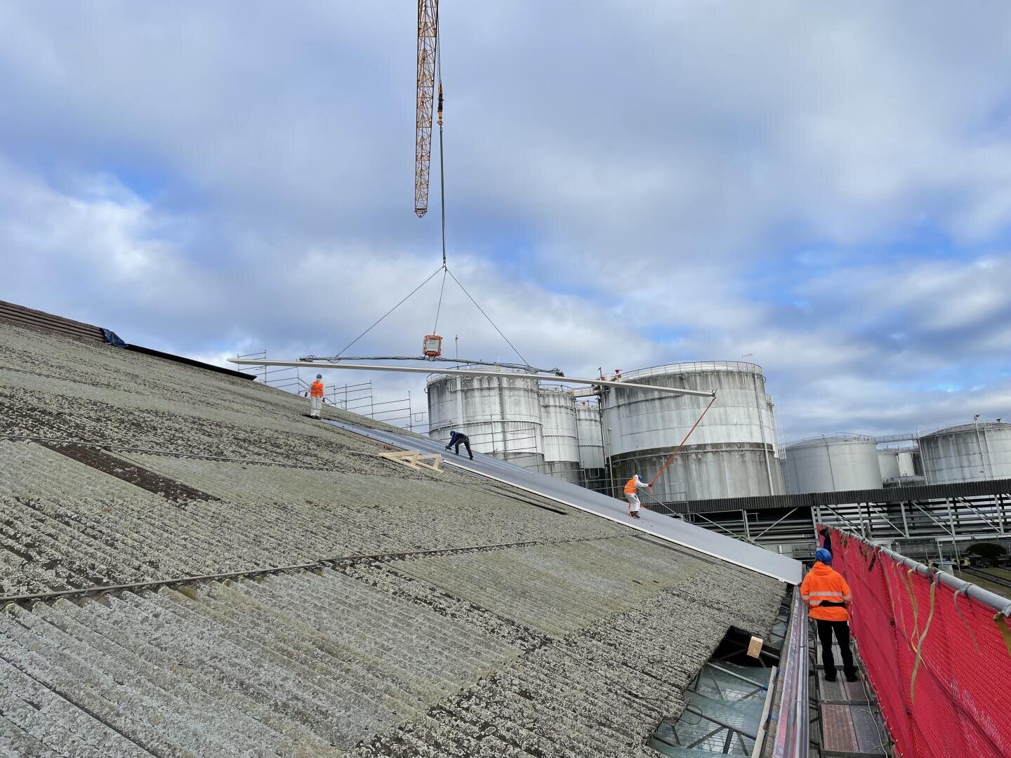 Mehrere Arbeiter in orangefarbenen Sicherheitsjacken montieren mit einem Kran große Platten auf einem schrägen Industriedach, mit zylindrischen Lagertanks und einem bewölkten Himmel im Hintergrund. Rote Sicherheitsnetze säumen den Dachrand.