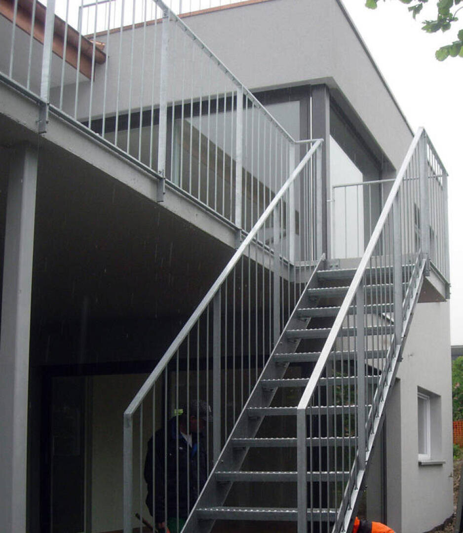 Eine Metalltreppe mit Geländer führt in den zweiten Stock eines modernen grauen Gebäudes mit großen Fenstern und einem Flachdach.