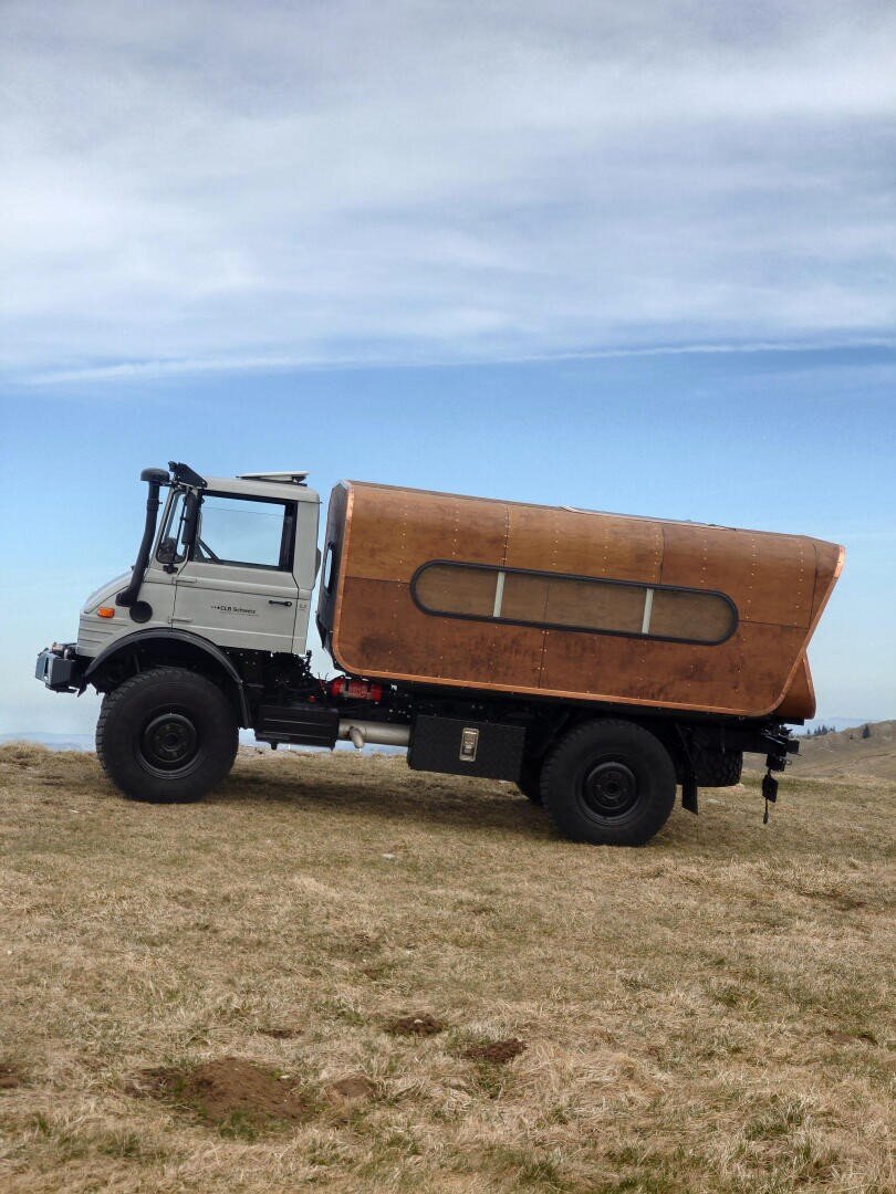 Ein weißer Unimog mit einem großen, speziell angefertigten Wohnmobil aus Holz, das auf trockenem Gras unter einem teilweise bewölkten Himmel geparkt ist.