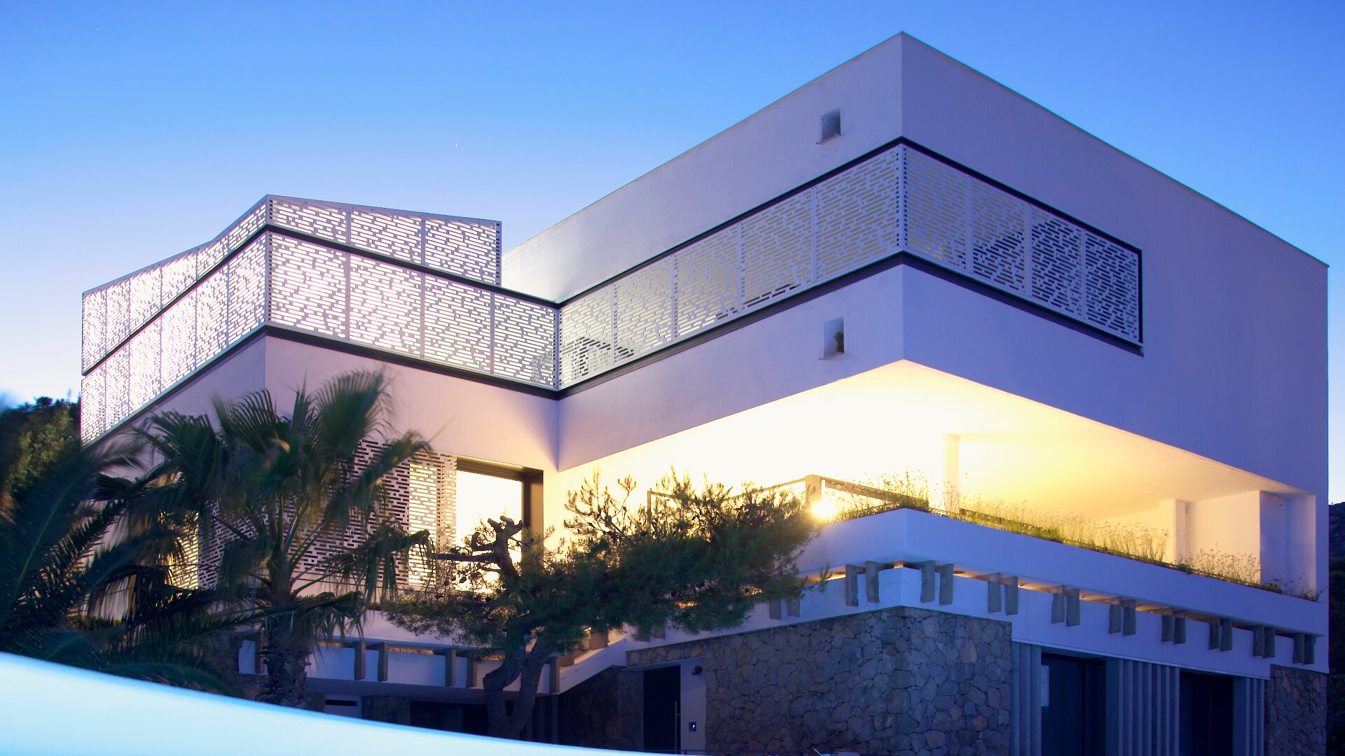 Ein modernes zweistöckiges Haus mit großen geometrischen Formen, heller Außenbeleuchtung, dekorativen Sichtschutzwänden und Steinakzenten, umgeben von tropischen Pflanzen, fotografiert in der Abenddämmerung bei klarem Himmel.