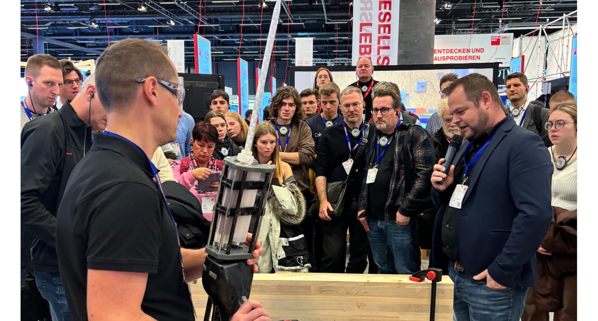 Eine Gruppe von Menschen versammelt sich um einen Redner, der ein mechanisches Gerät hält, während eine andere Person bei einer Veranstaltung oder Messe in einem Gebäude in ein Mikrofon spricht. Im Hintergrund sind Banner und Displays zu sehen.
