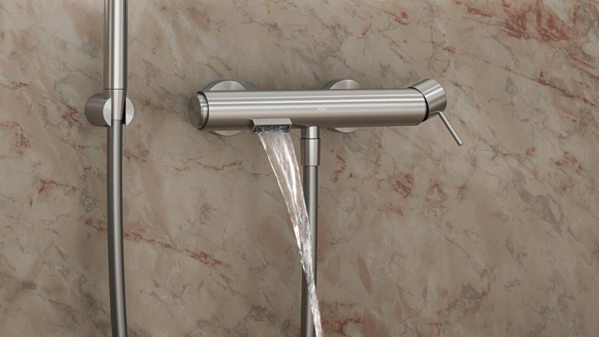 Un robinet de douche moderne en acier inoxydable libère un jet d'eau sur un mur carrelé marbré beige et marron, avec une pomme de douche et une poignée visibles sur le côté gauche.