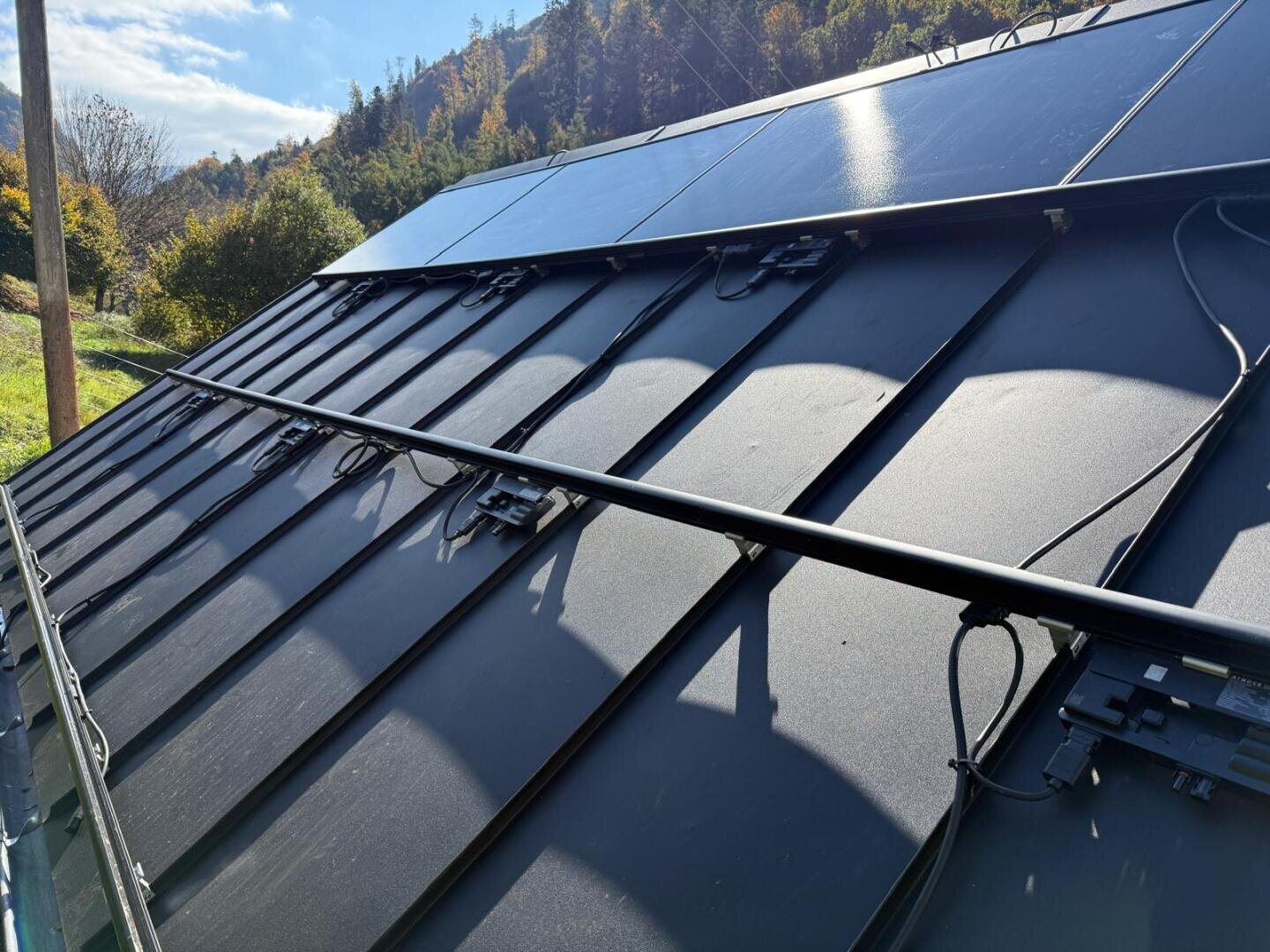 Auf einem Metalldach installierte Solarmodule mit sichtbaren Kabeln und elektronischen Bauteilen darunter, vor einem Hintergrund von Bäumen und einem teilweise bewölkten Himmel.