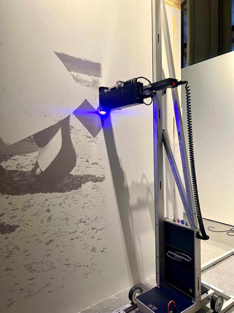 Ein Wanddrucker strahlt ein blaues Licht aus, während er ein Graustufenbild auf eine große vertikale Fläche in einem Gebäude druckt. Das Bild an der Wand scheint einen Teil einer abstrakten Szene oder einer Szene im Freien zu zeigen.