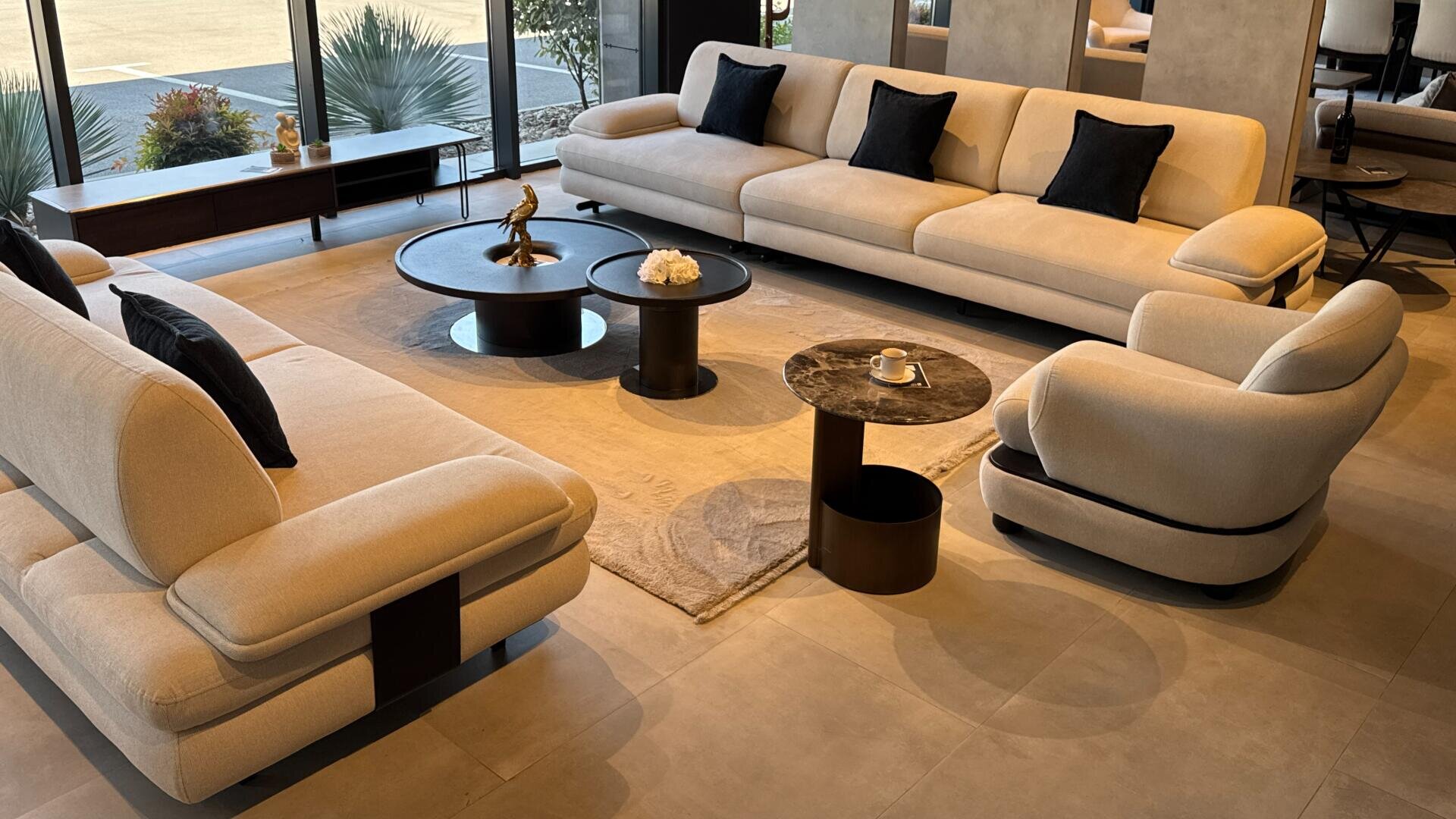Modernes Wohnzimmer mit zwei beigen Sofas, einem passenden Sessel, drei runden Couchtischen, schwarzen Polstern und einem beigen Teppich. Große Fenster geben den Blick auf Pflanzen und den Außenbereich frei. Der Raum hat ein minimalistisches, elegantes Design.