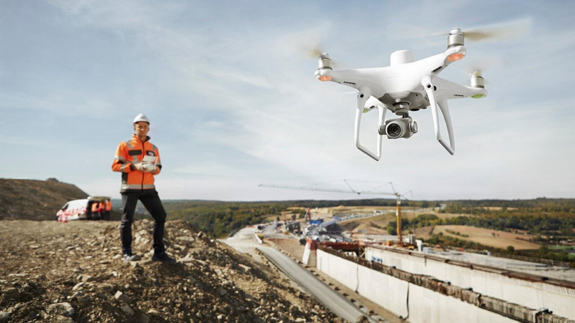 Une personne en tenue de sécurité pilote un drone blanc au-dessus d'un chantier de construction avec des grues, des véhicules et une route ou un pont partiellement construit dans un paysage rural.