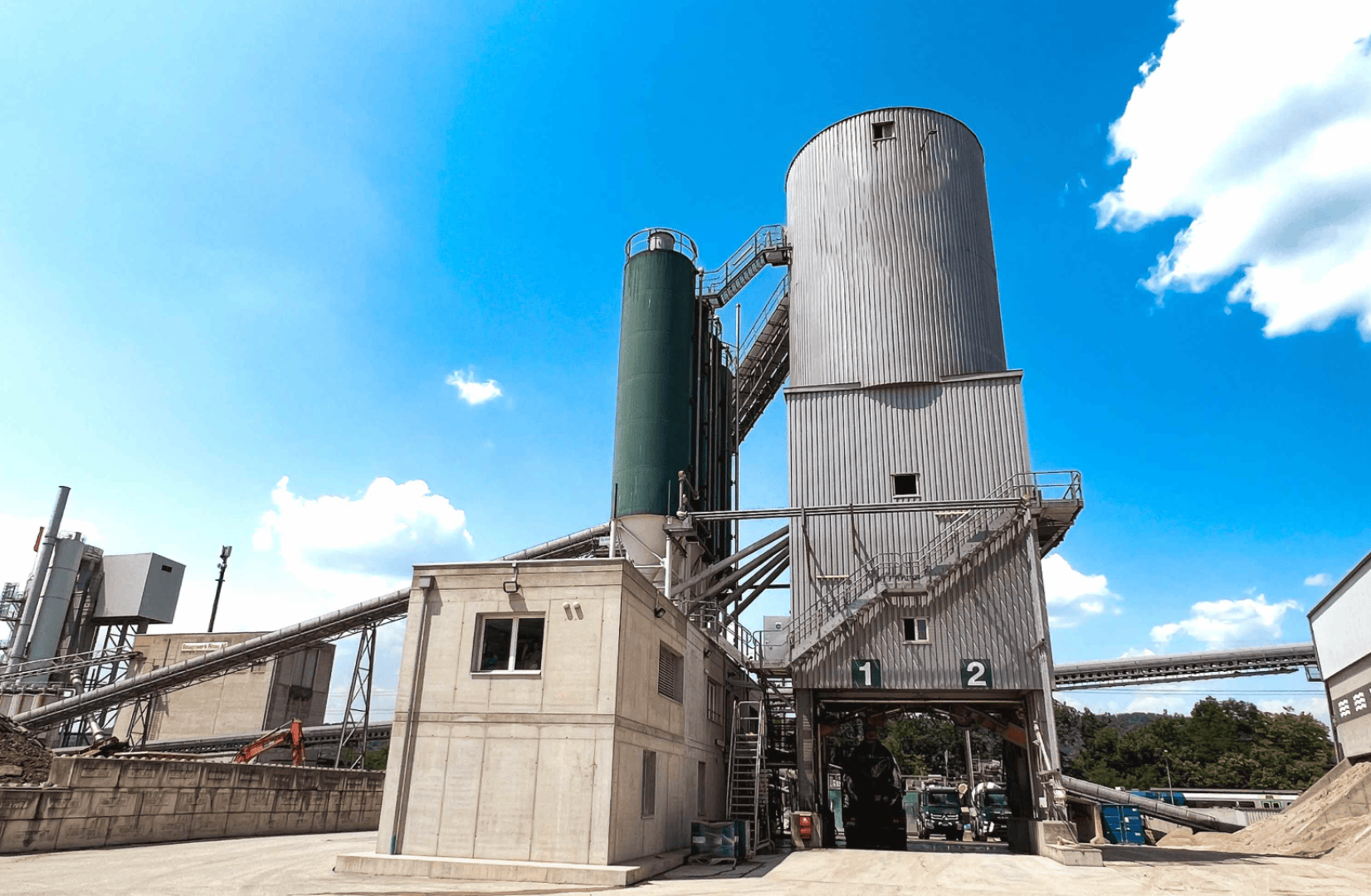 Installation industrielle avec de hauts silos cylindriques, des escaliers métalliques et un petit bâtiment en béton à l'avant. Des convoyeurs relient les structures. Ciel bleu vif avec des nuages épars en arrière-plan.