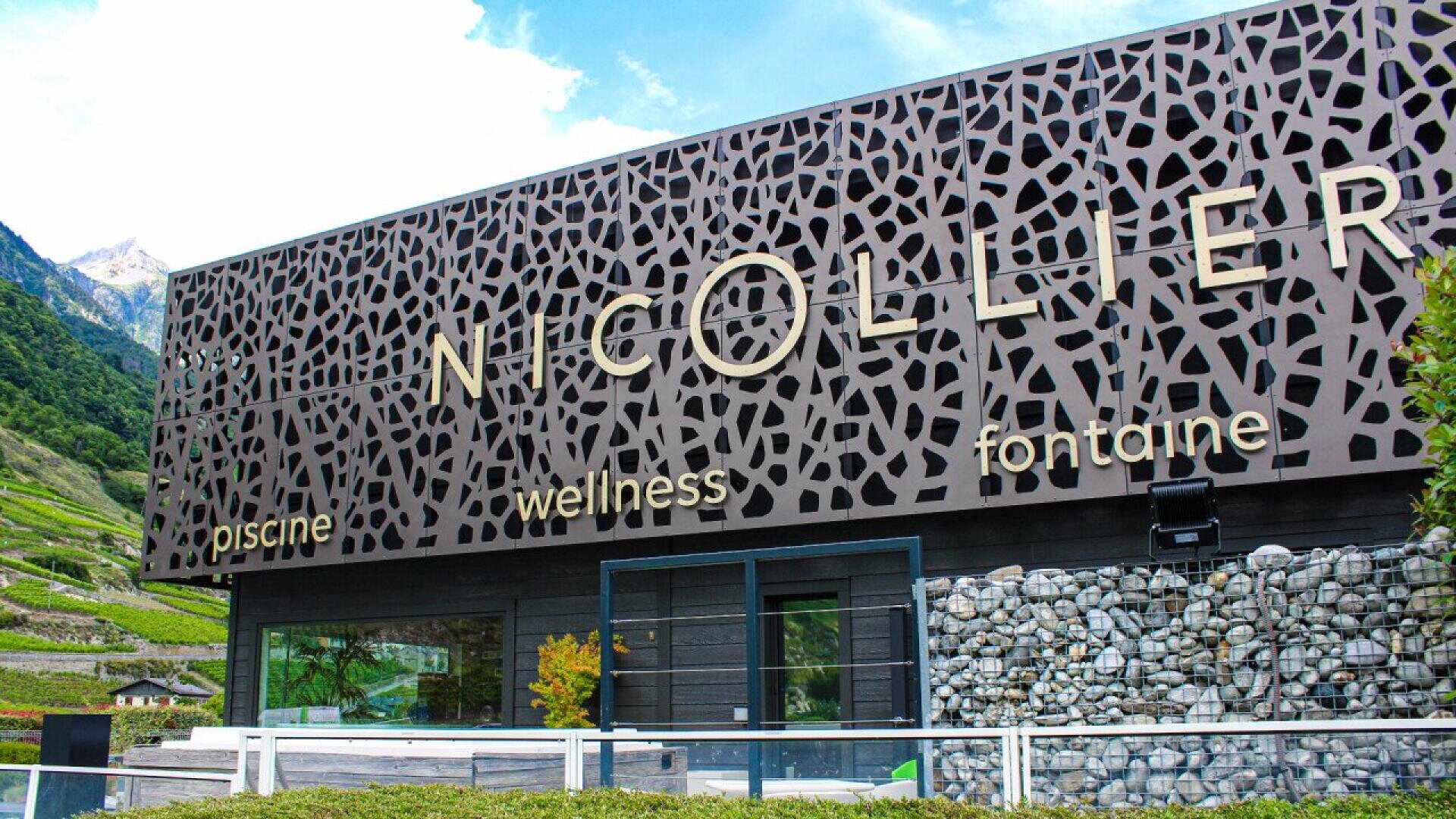 Un bâtiment moderne avec une façade décorative perforée affiche les mots NICOLLIER piscine wellness fontaine. Des collines vertes en terrasse et des montagnes sont visibles à l'arrière-plan sous un ciel bleu.