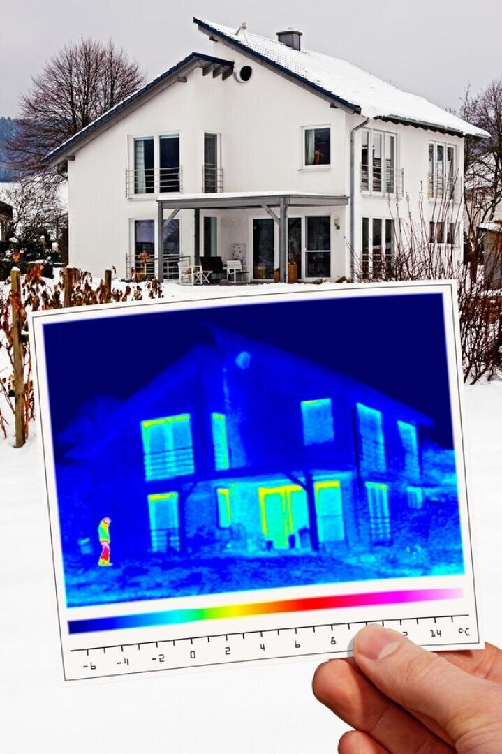 Eine Person hält ein Wärmebild vor ein weißes Haus im Winter, das Wärmeverlustbereiche in leuchtendem Gelb und Orange und kühlere Bereiche in Blau und Grün zeigt. Der Boden ist mit Schnee bedeckt, und Bäume umgeben das Haus.