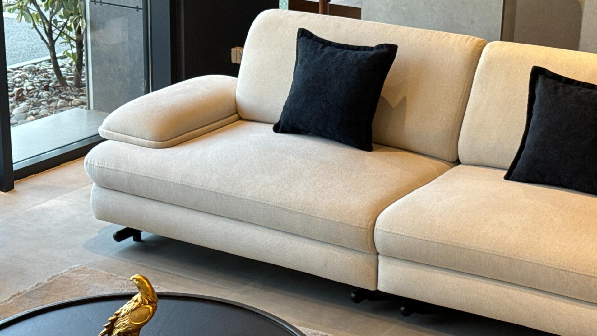 Ein modernes beiges Sofa mit zwei schwarzen Kissen steht in einem stilvollen Wohnzimmer neben großen Fenstern. Vor dem Sofa steht ein runder schwarzer Couchtisch mit einem goldenen Deko-Objekt.