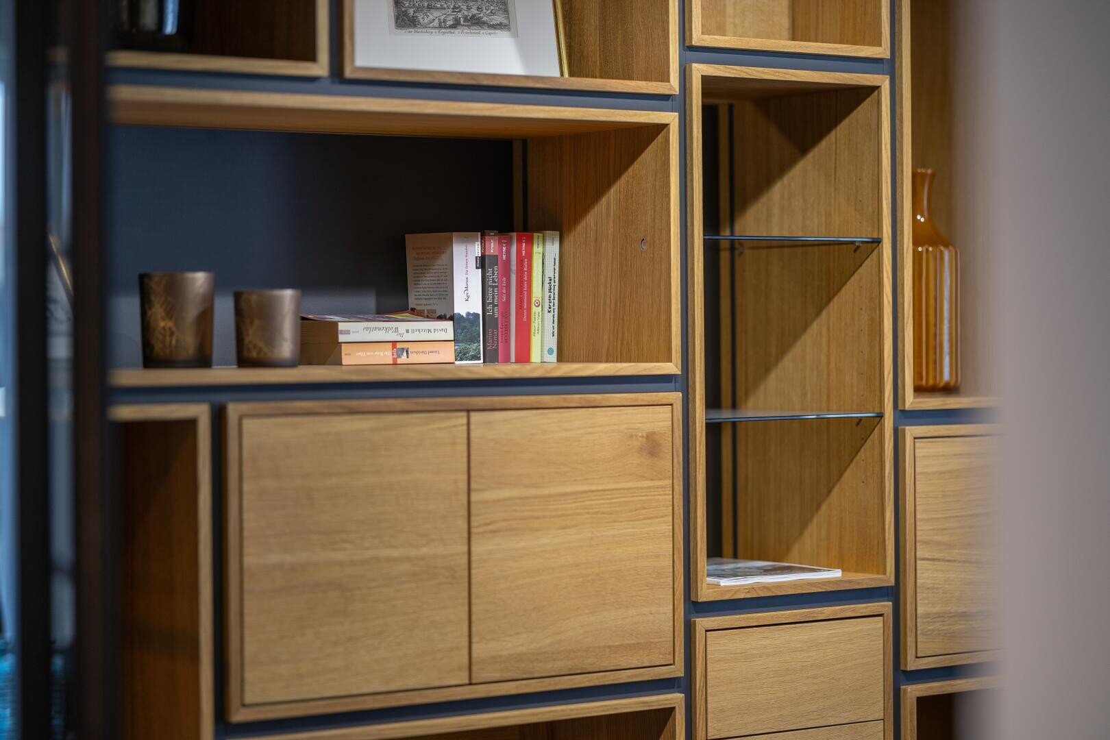Ein modernes Holzregal mit offenen Fächern, ein paar gestapelte Bücher, zwei braune Tassen, eine orangefarbene Glasvase und gerahmte Kunstwerke, die in einem minimalistischen Stil ordentlich angeordnet sind.