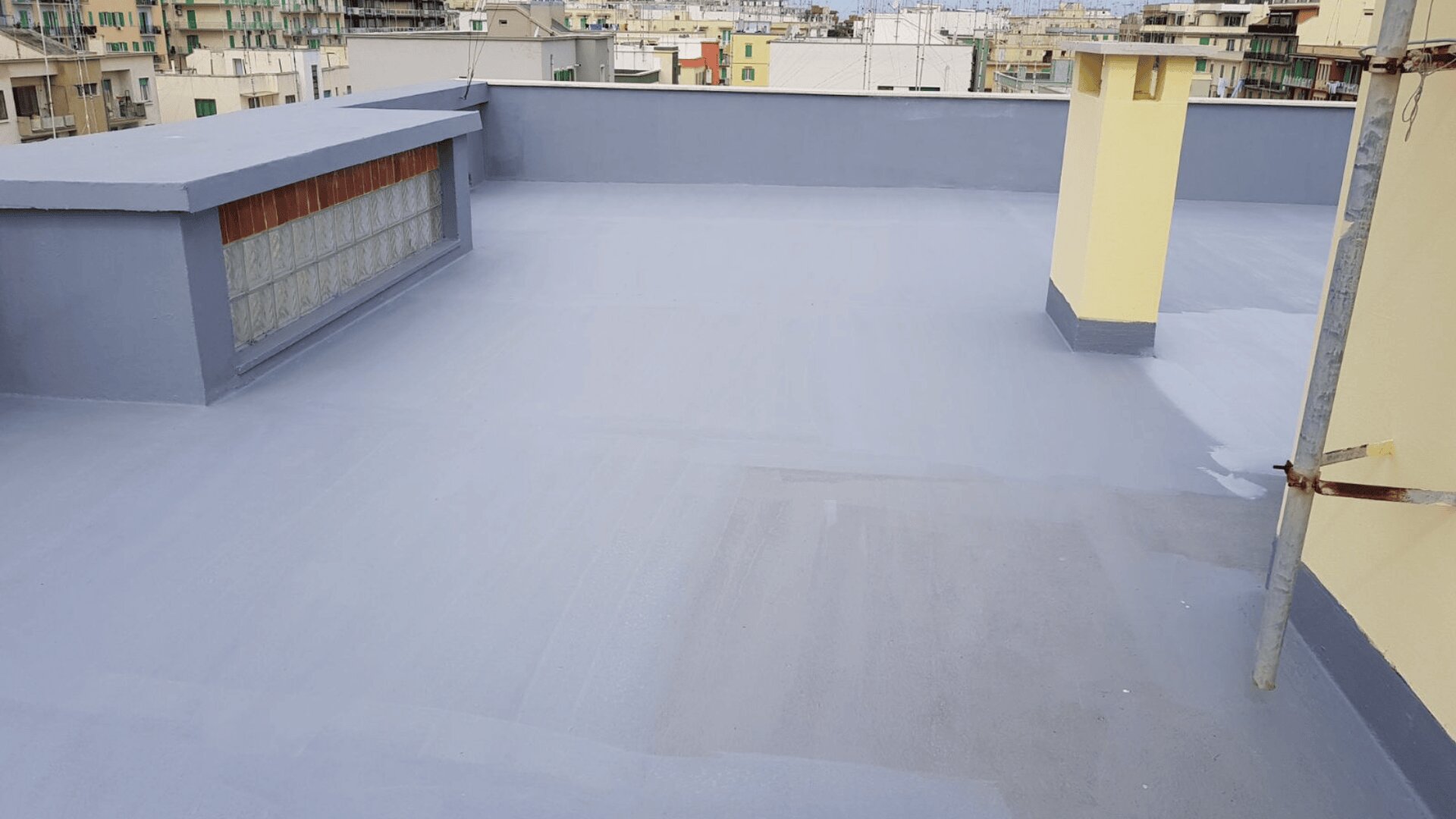 Toit plat recouvert d'un revêtement lisse et imperméable de couleur grise, bordé de murets. Des bâtiments avec balcons sont visibles à l'arrière-plan sous un ciel partiellement nuageux.